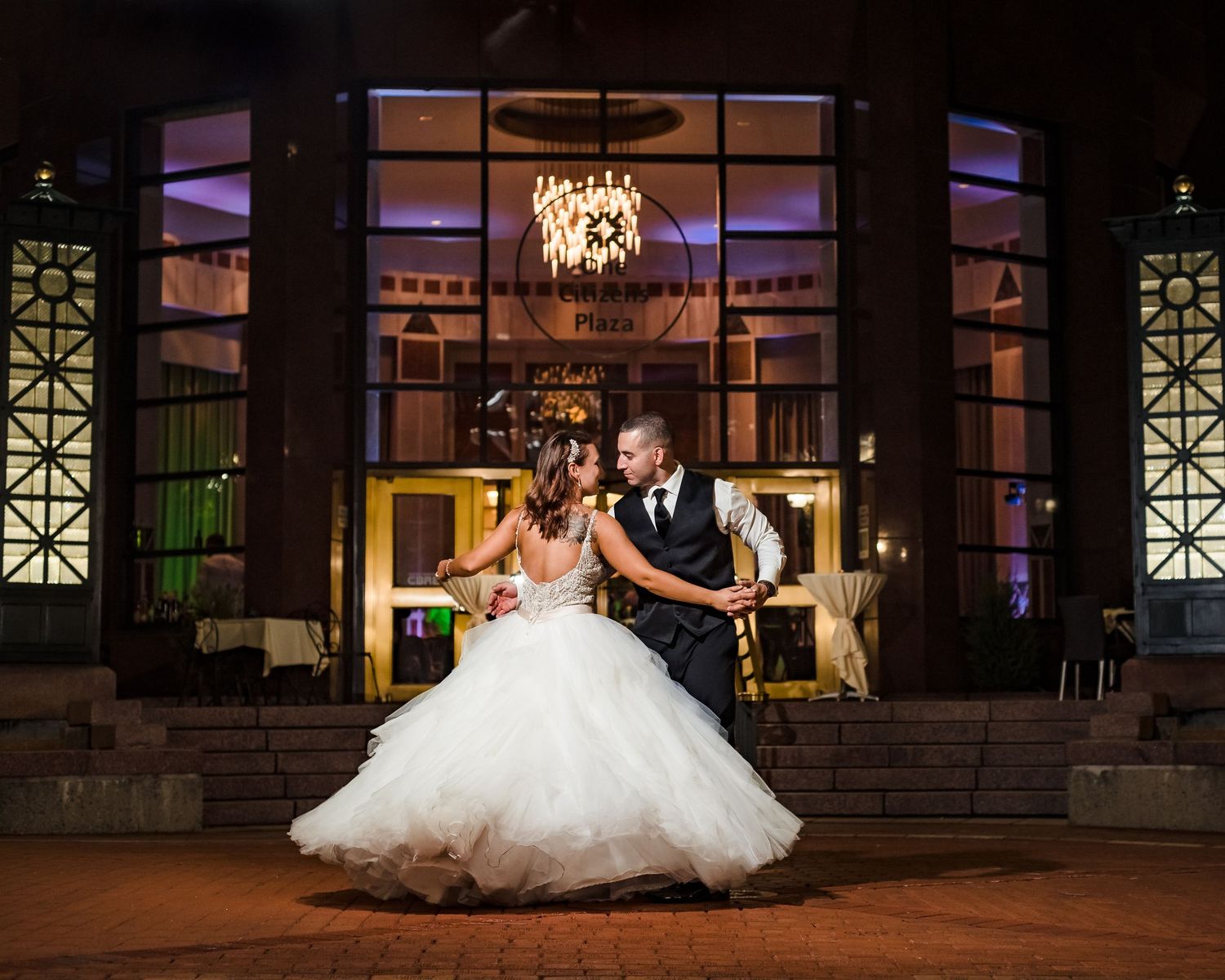 Newlywed couple dances together in an elegant ballroom with ornate chandeliers and decorative windows.