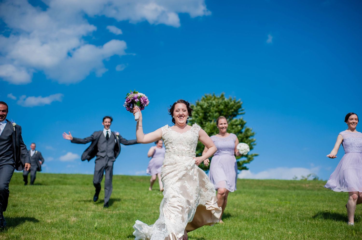 Wedding party having fun running across green hillside under blue sky, photographed by a Rhode Island wedding photographer