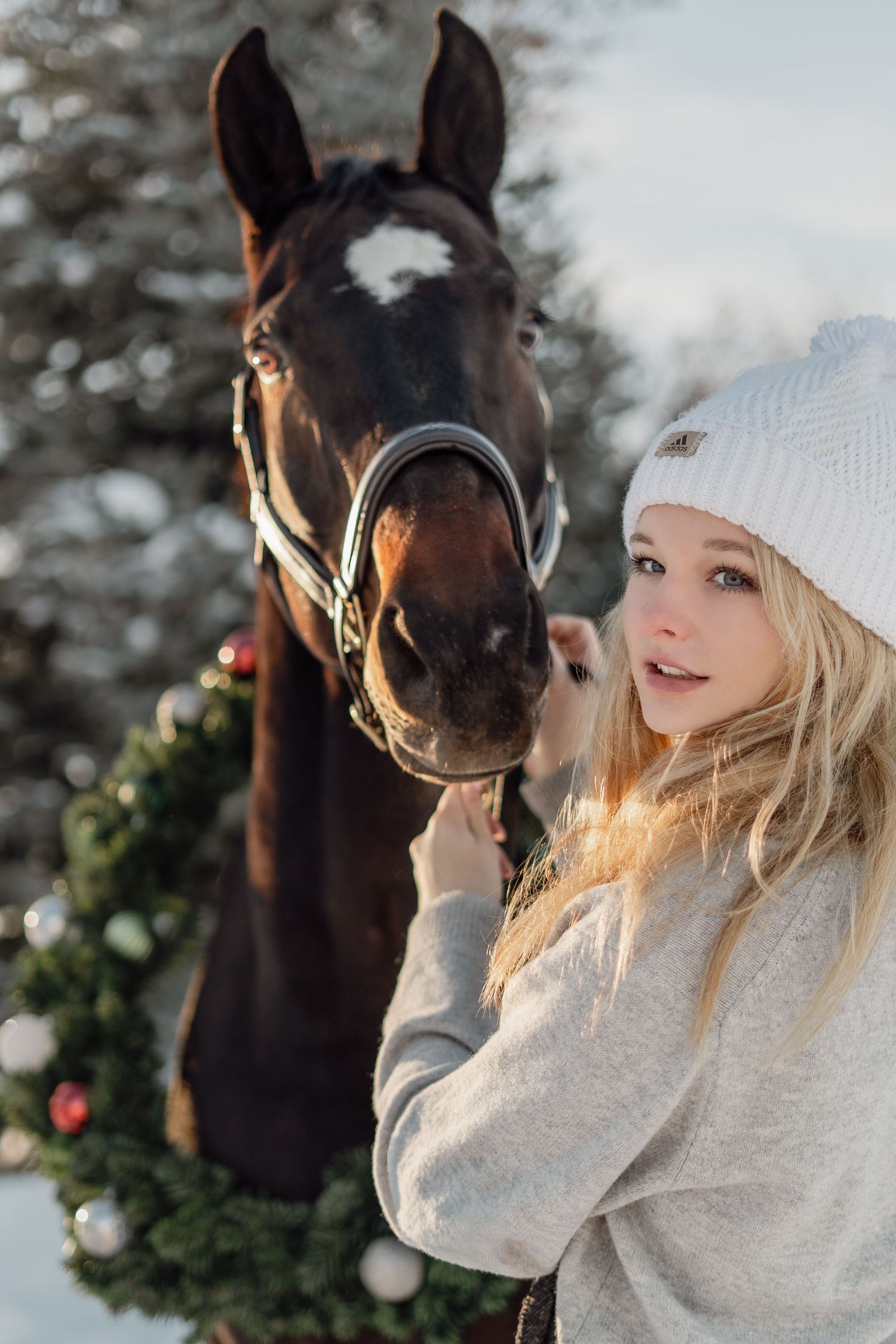 Young blonde girl in white winter hat stands beside a dark brown horse with Christmas wreath in snowy setting.