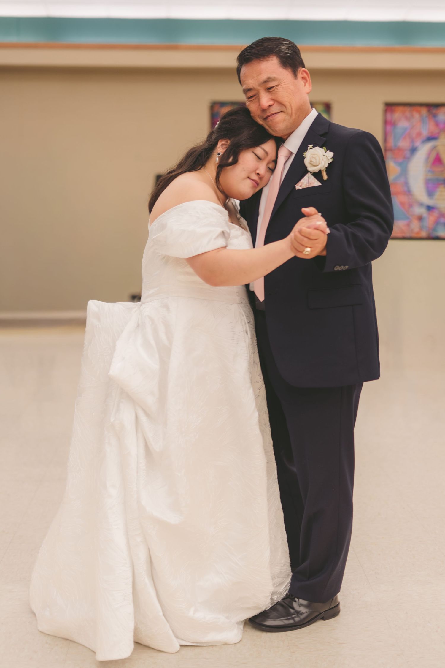 Bride in white off-shoulder gown shares emotional father-daughter dance with man in dark suit at wedding reception.