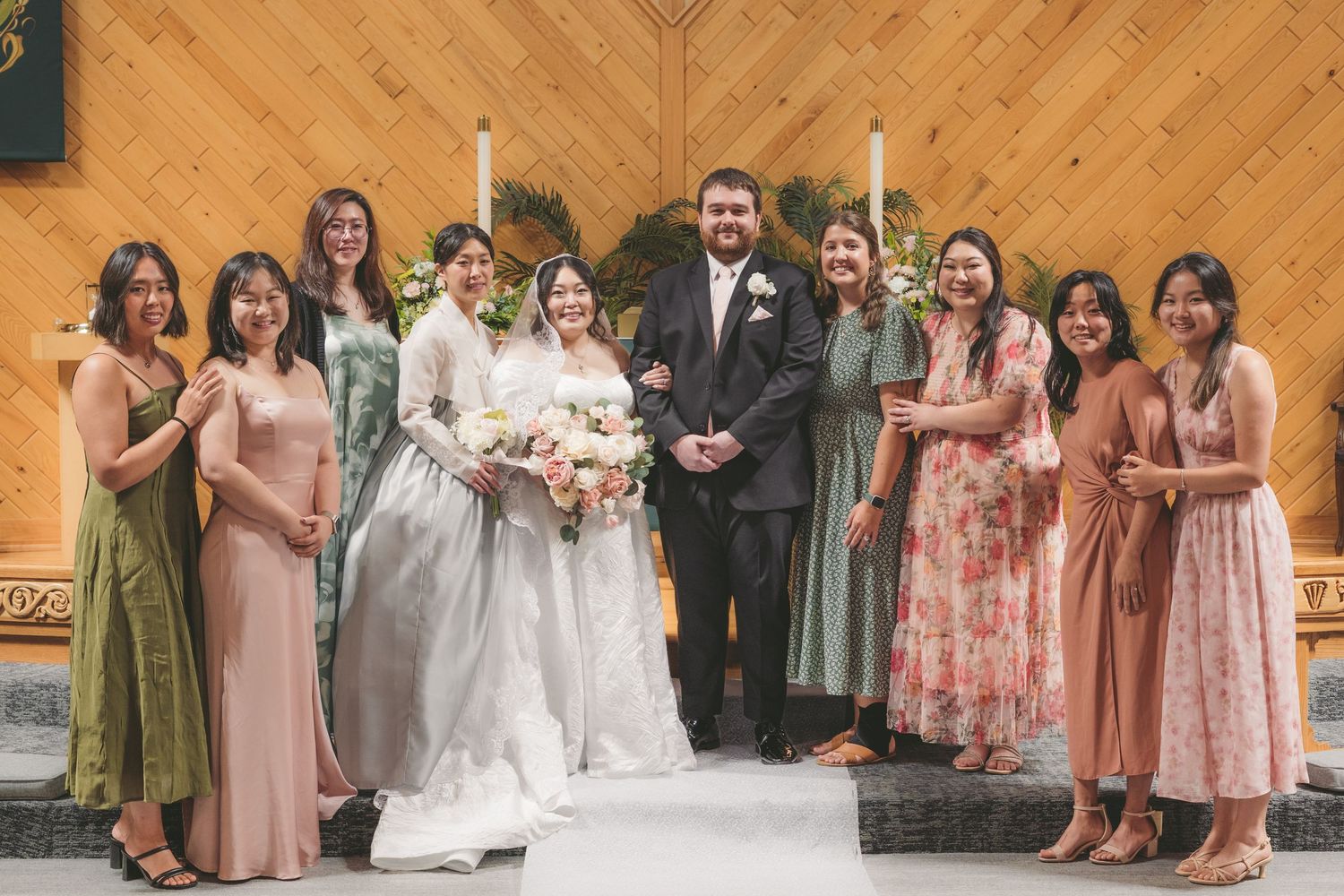 Wedding party posing in a wooden chapel, bride in white gown with groom and bridesmaids in sage green and pink dresses.