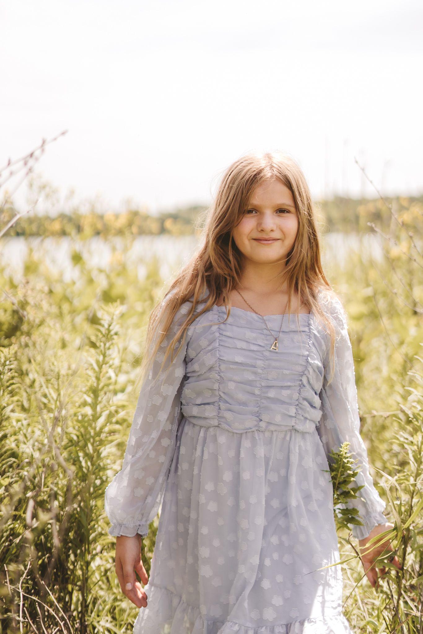 Young girl in a light blue polka dot dress standing in a sunny green field near a lake, smiling softly.