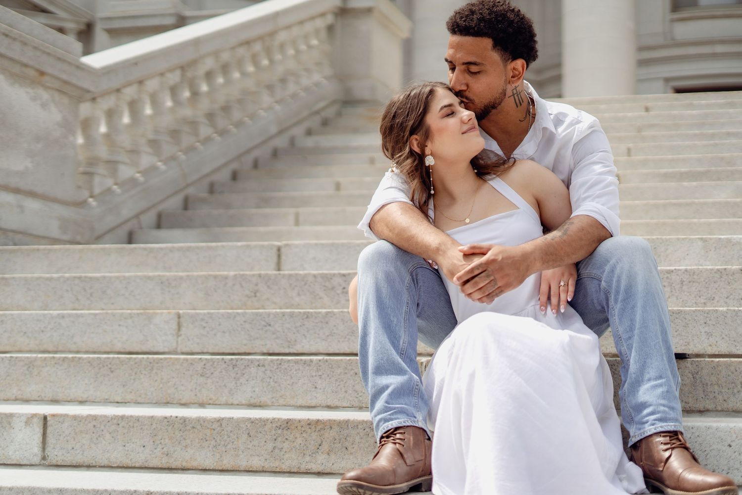 Romantic couple sitting on stone steps, man kissing woman's head, wearing casual white outfits in an elegant setting.