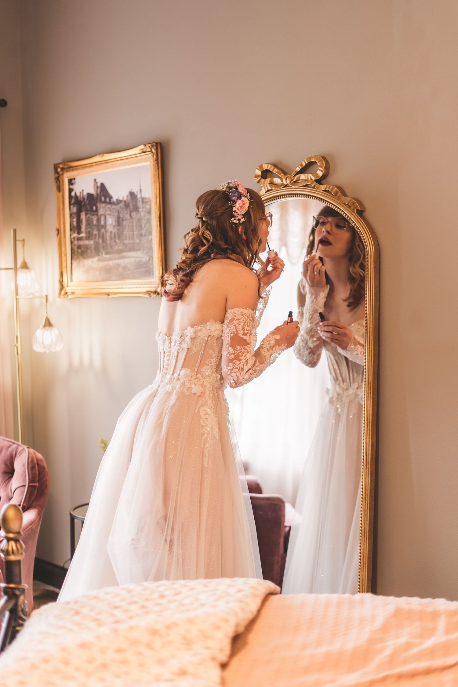 A bride in a lace off-shoulder wedding gown applies lipstick while gazing into an ornate gold mirror.