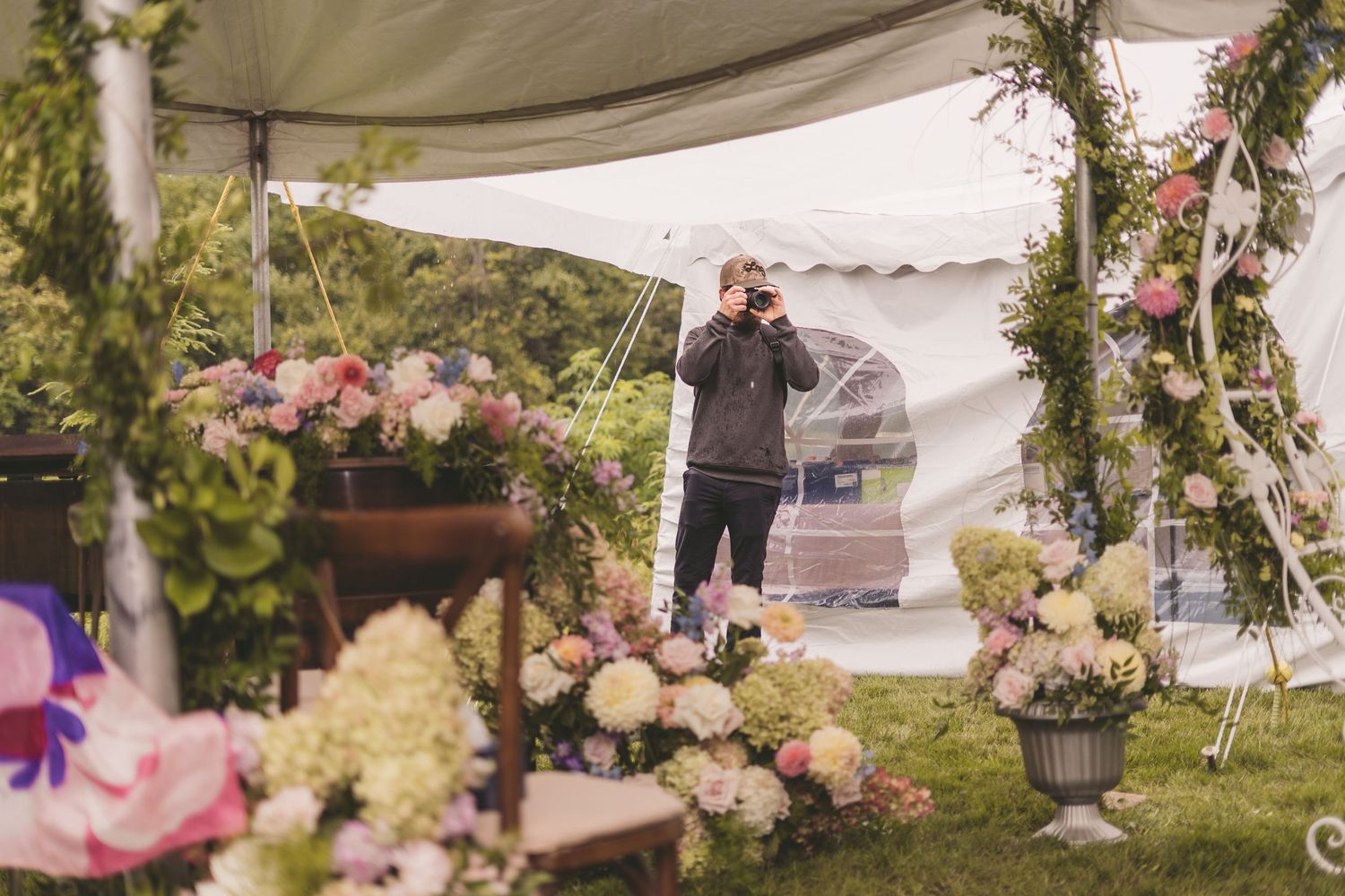 Photographer capturing floral wedding decorations with pink hydrangeas and roses under a white tent on a green lawn.