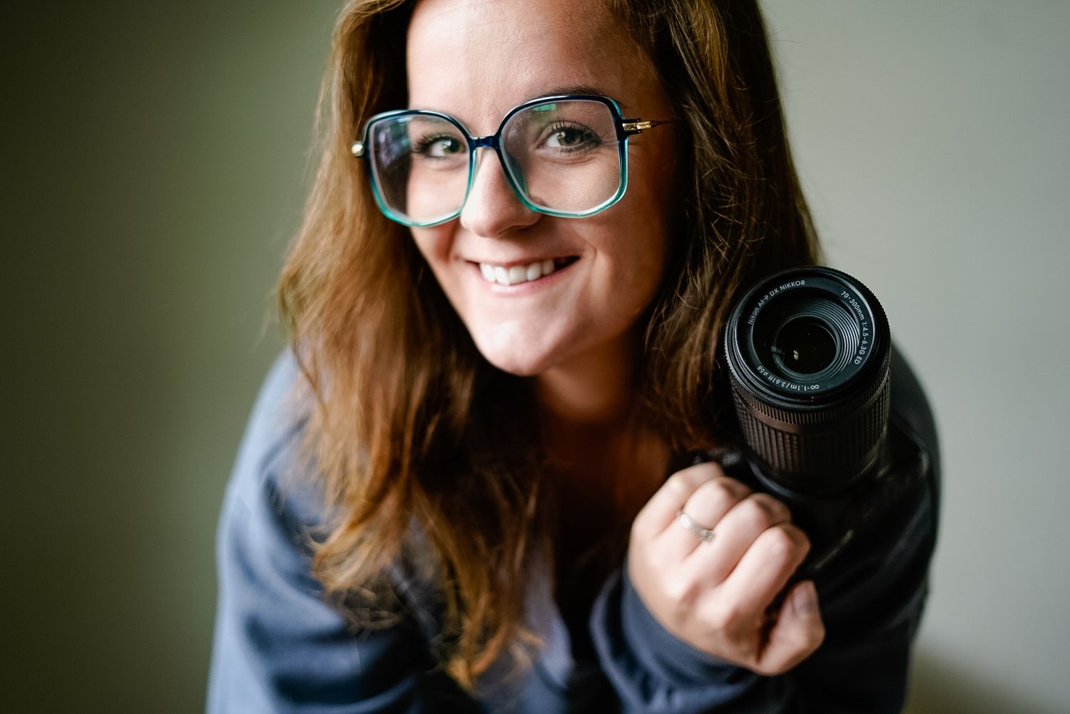 A photographer holds up a camera lens while wearing glasses and a blue sweater.