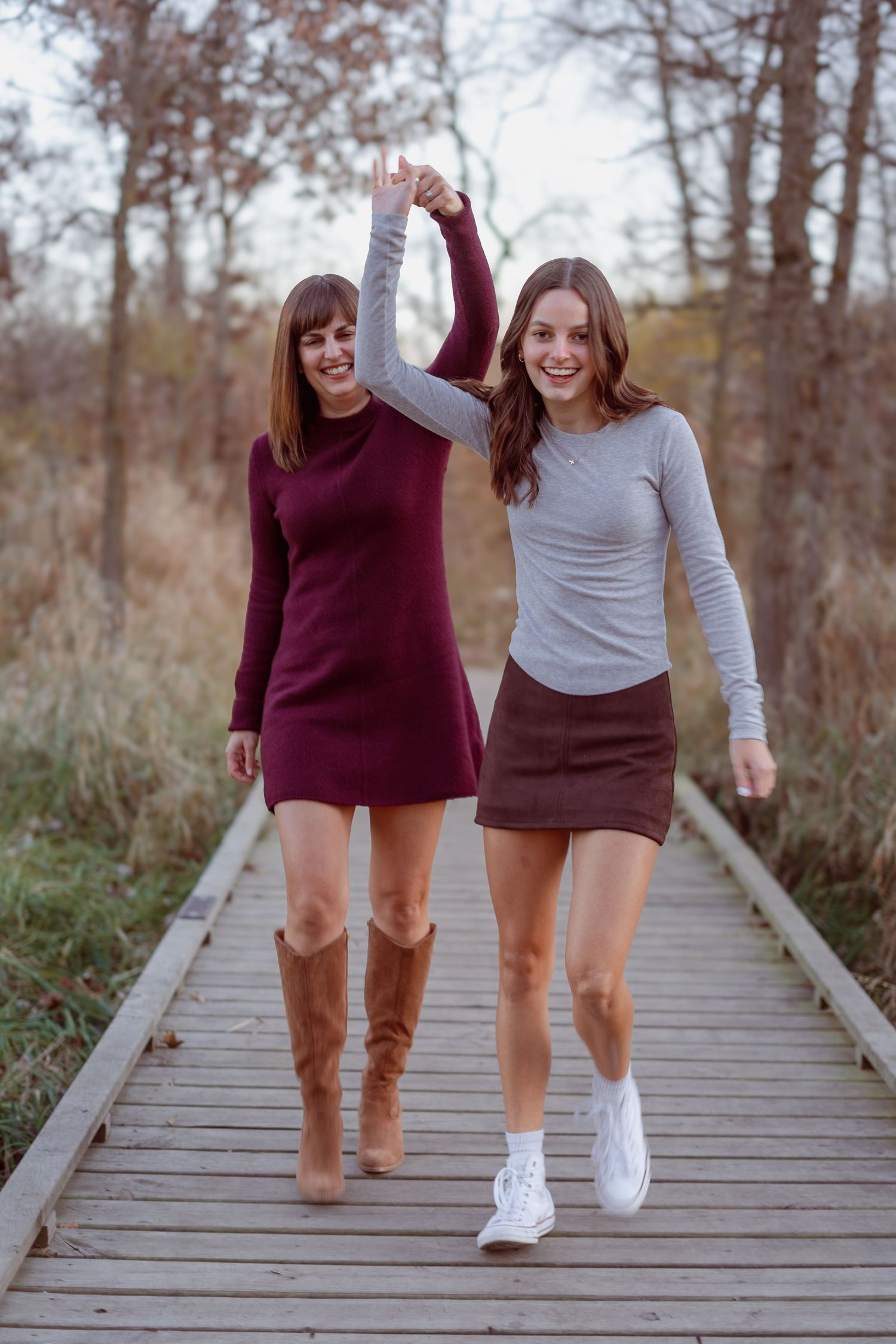 Two smiling women holding hands walk on a wooden boardwalk path in autumn, wearing mini dresses and boots.