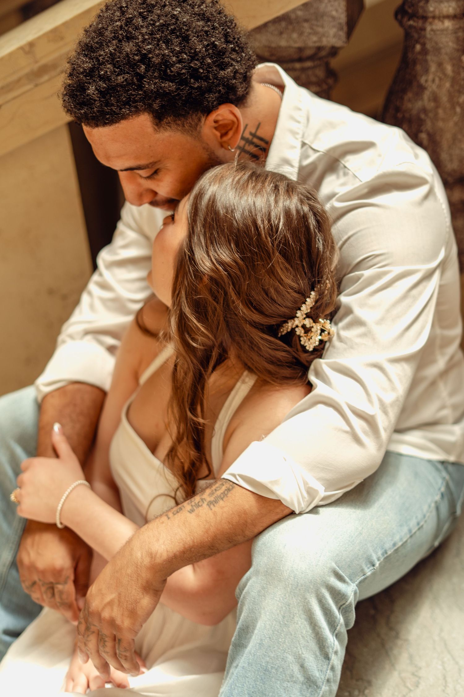 Romantic couple sitting closely together on steps, man embracing woman with brown hair accessory from behind.