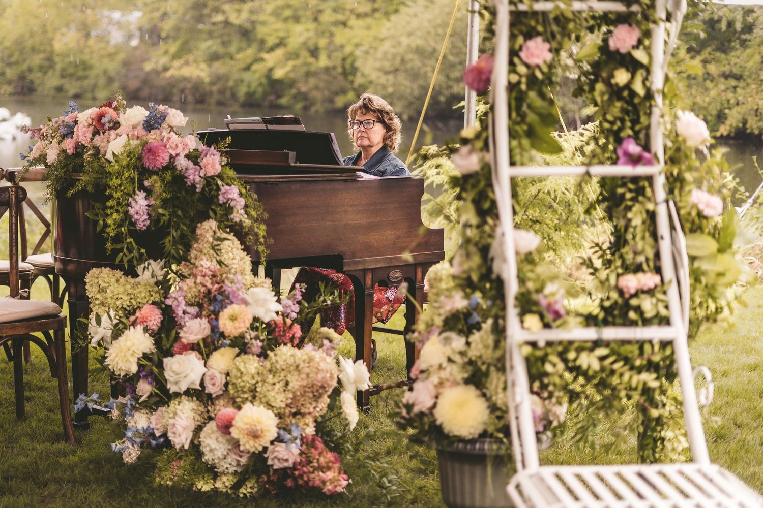 Outdoor wedding pianist surrounded by lush pink and white floral arrangements in a garden setting.