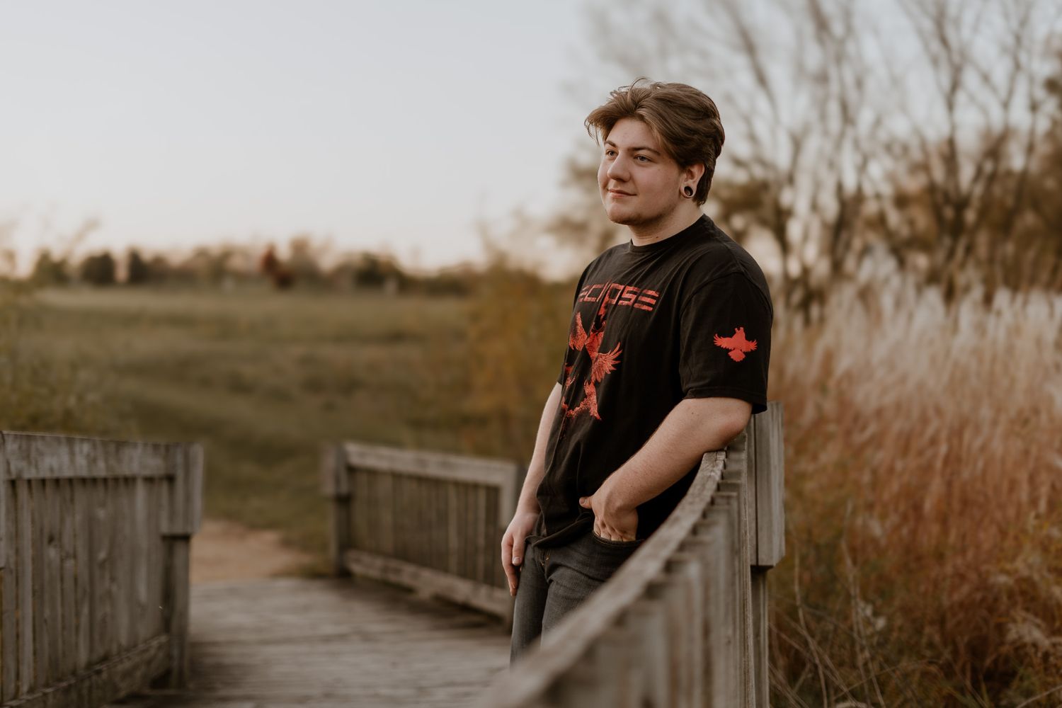 A person in a black t-shirt stands thoughtfully on a wooden boardwalk at sunset.