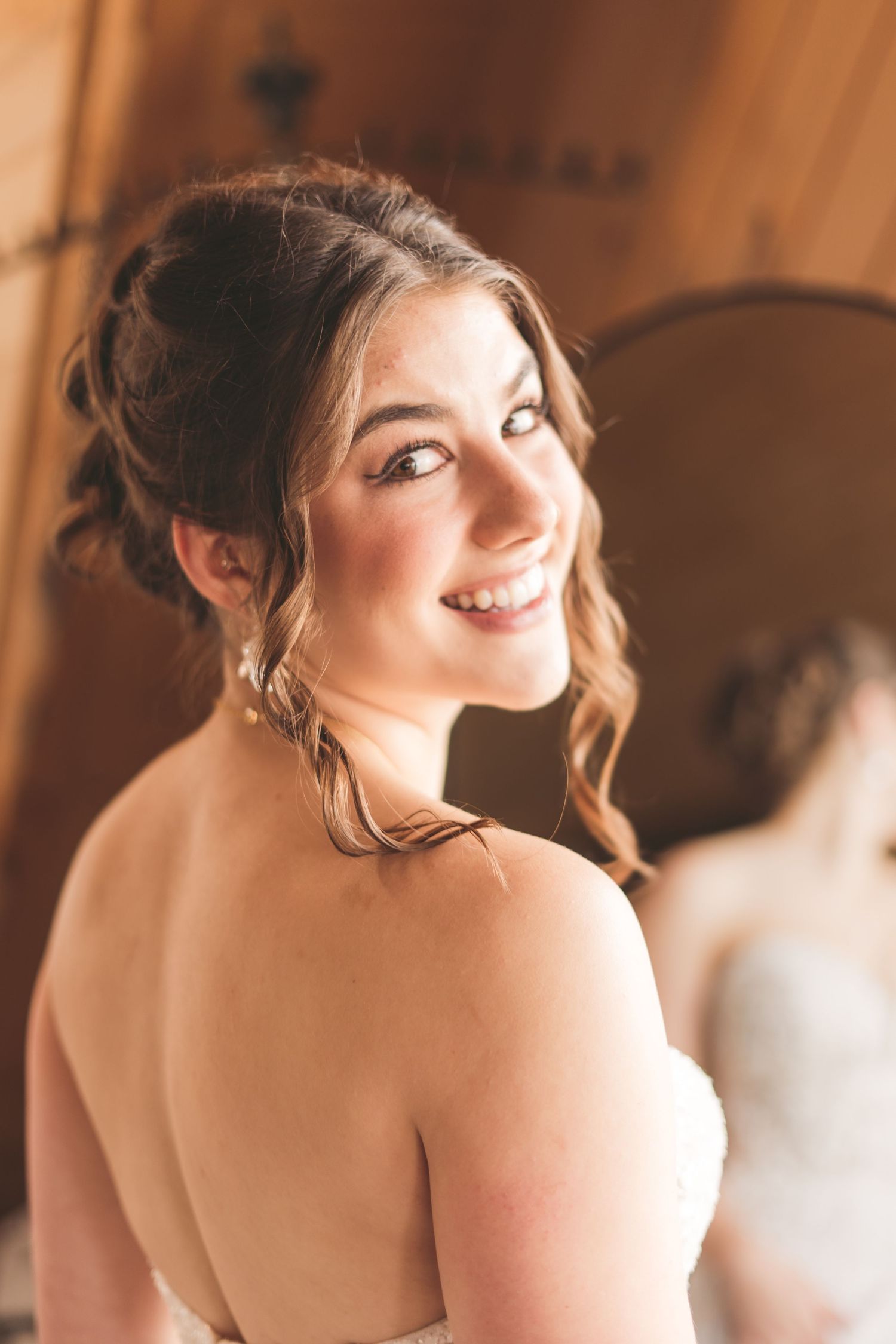 Smiling bride in strapless white gown with elegant updo hairstyle, glancing over her shoulder in warm light.