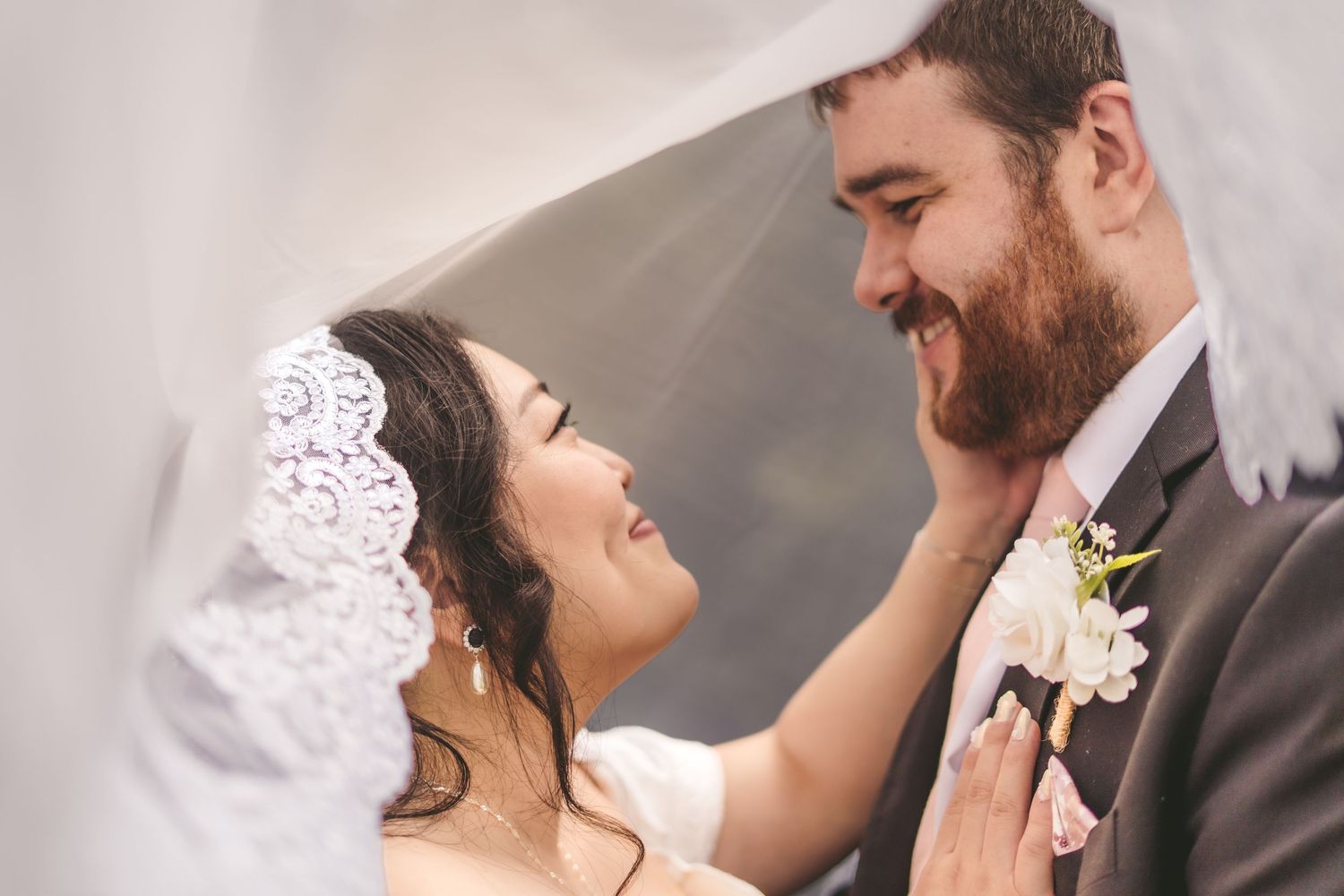 Bride and groom share a romantic moment under a wedding veil, smiling at each other on their wedding day.