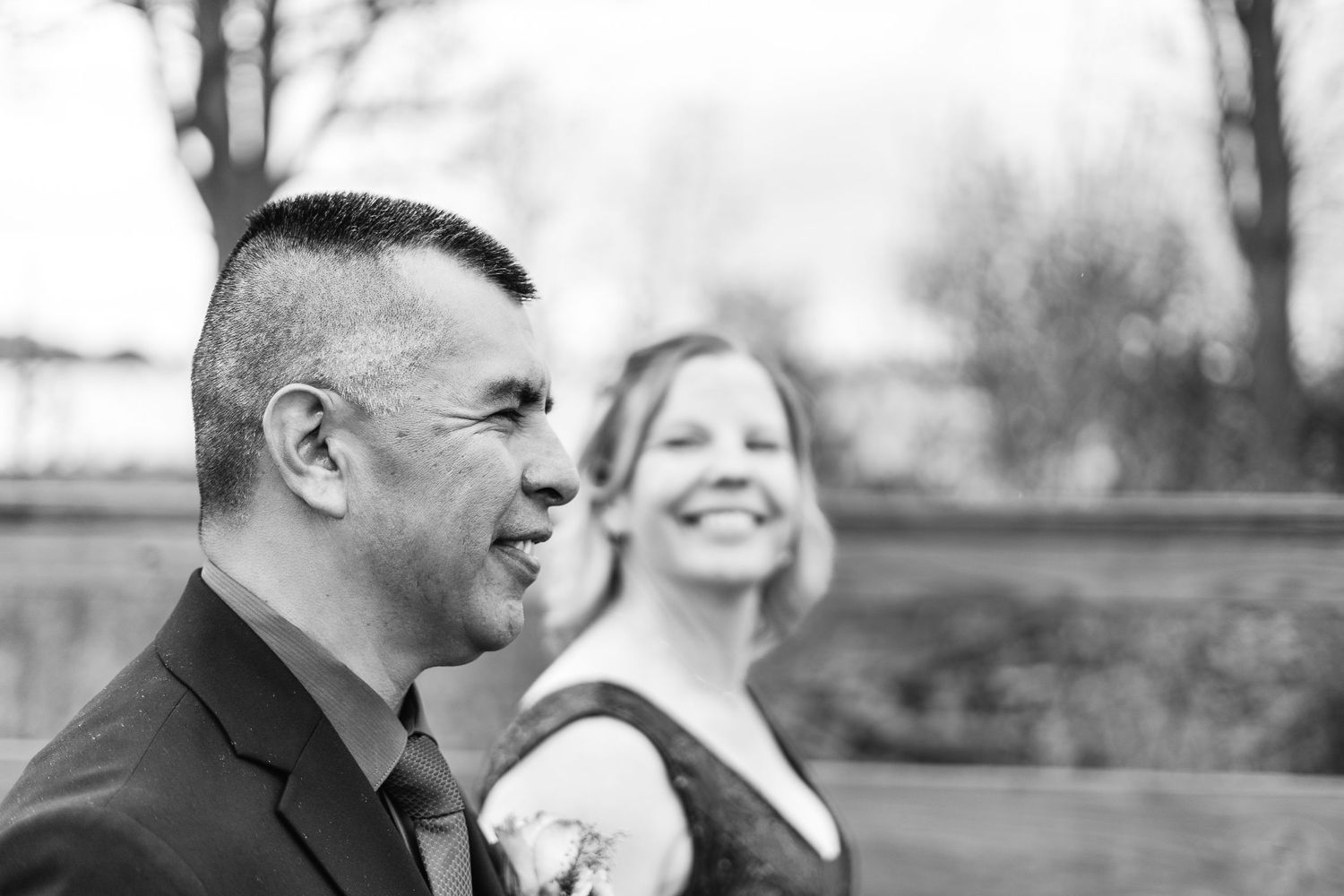 Black and white wedding photo of a smiling couple, groom in suit with boutonniere, bride laughing in background.