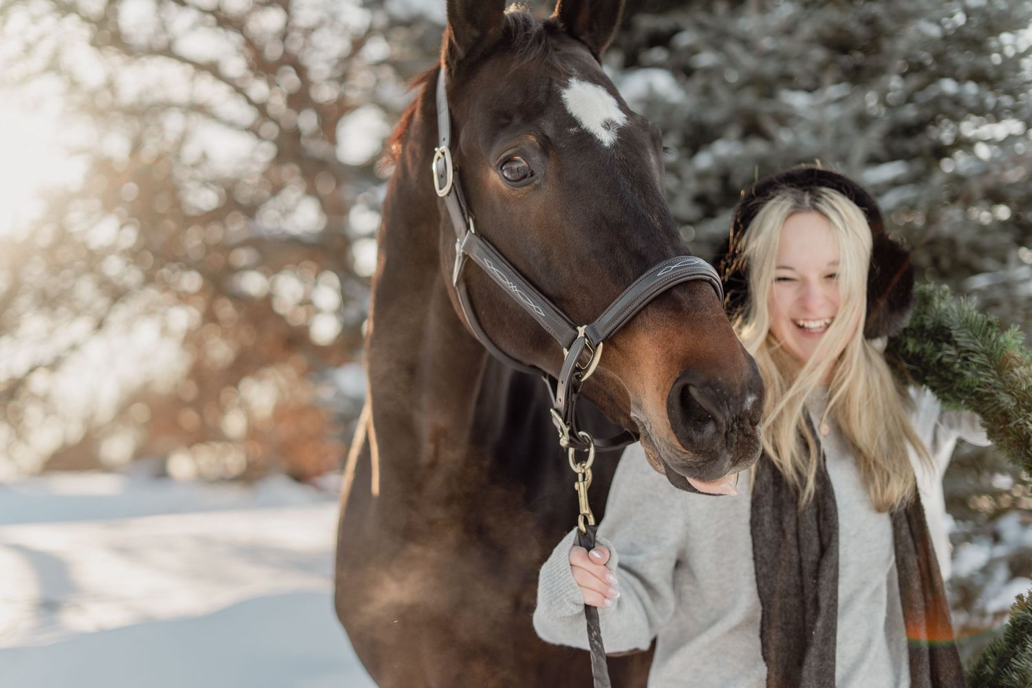 Smiling blonde woman with a dark bay horse in snowy winter landscape with sunlit trees.