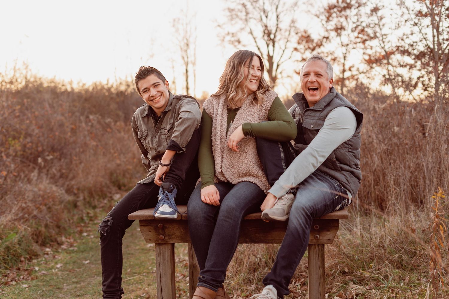 Three women laughing together on a wooden bench outdoors in autumn, enjoying a fun family photo session.