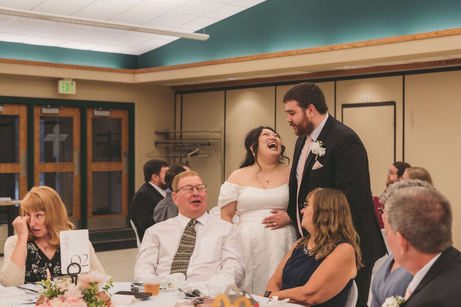 Bride and groom laughing joyfully with wedding guests during indoor reception celebration.