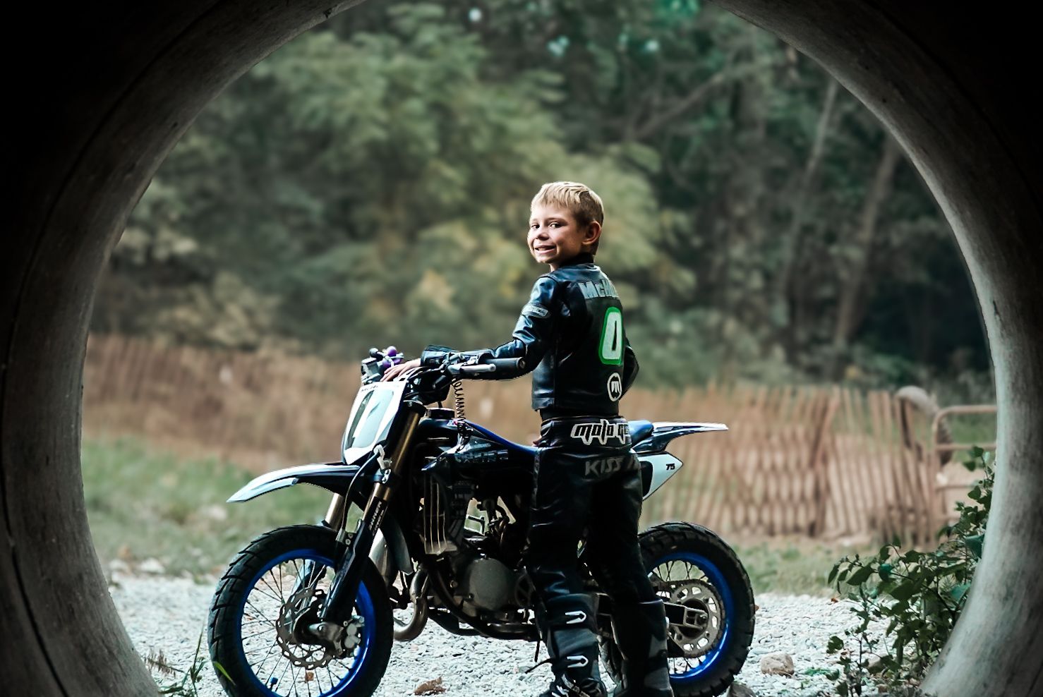 Young motocross rider in black leather jacket sitting on a blue dirt bike viewed through a concrete tunnel opening.