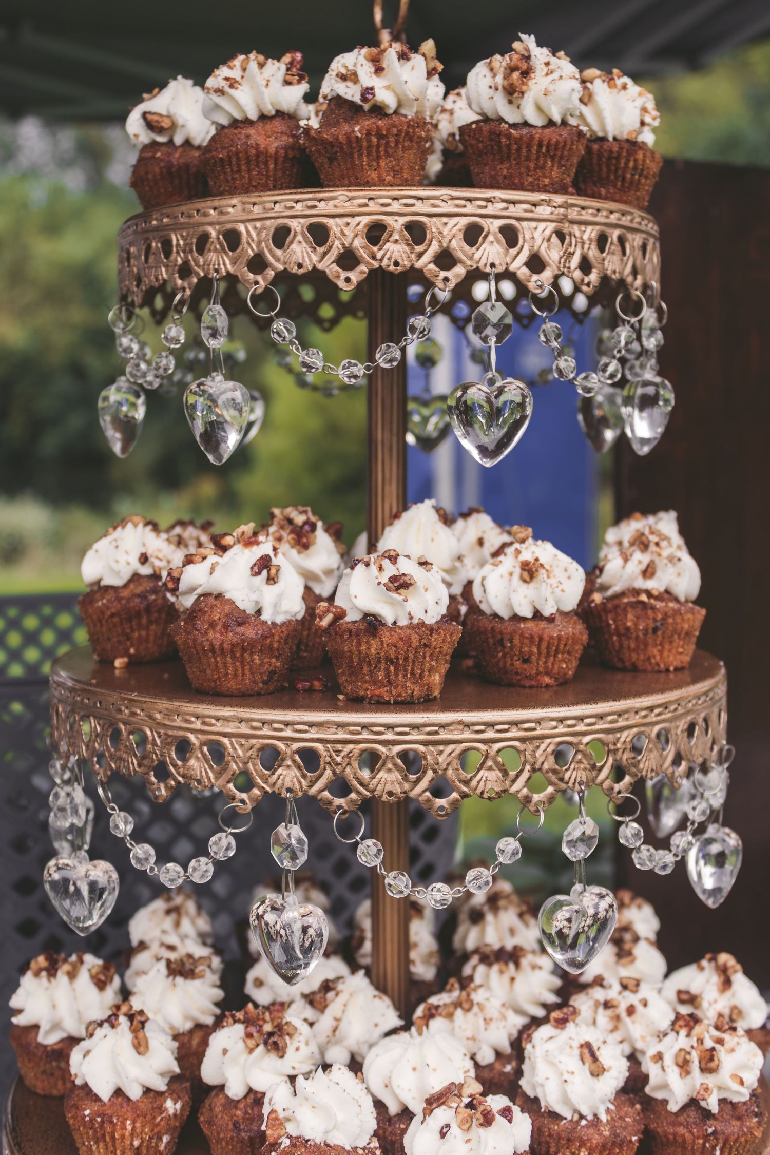 Three-tier ornate gold cupcake stand adorned with crystal heart pendants displaying chocolate cupcakes with white frosting.
