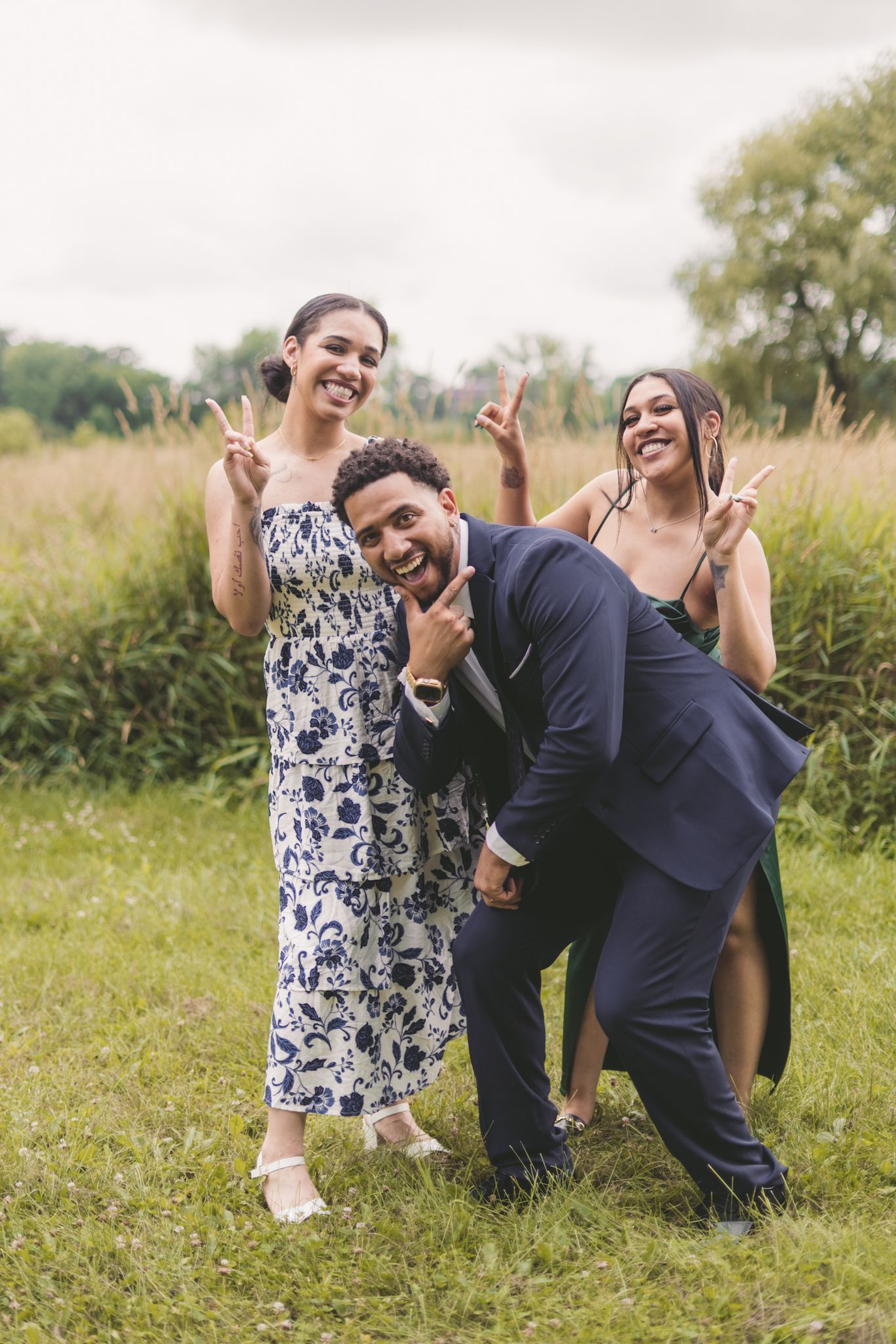 Three friends posing playfully outdoors, man in navy suit crouching while two women in elegant dresses flash peace signs.