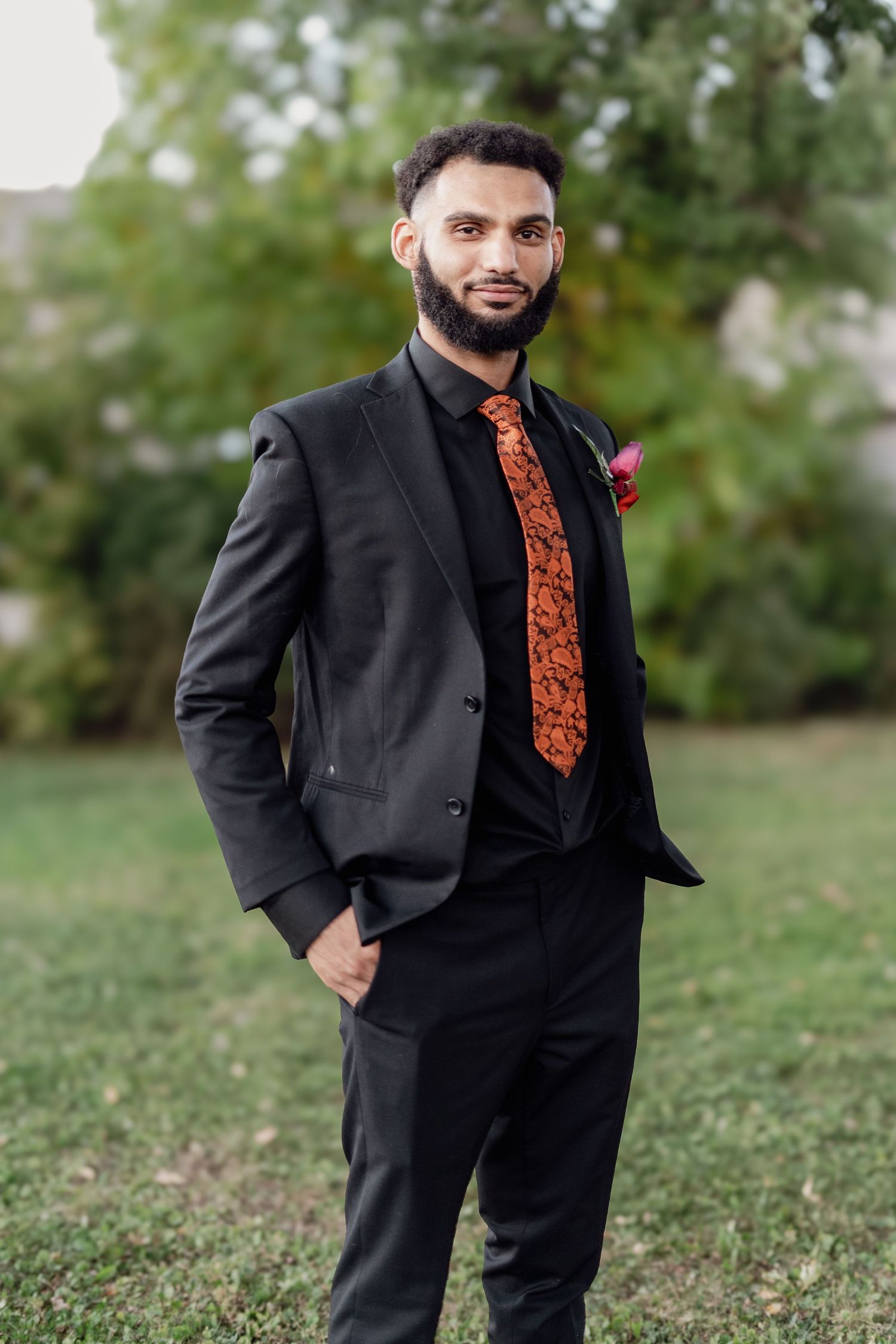 Person wearing a black suit with orange patterned tie poses outdoors on grassy lawn.