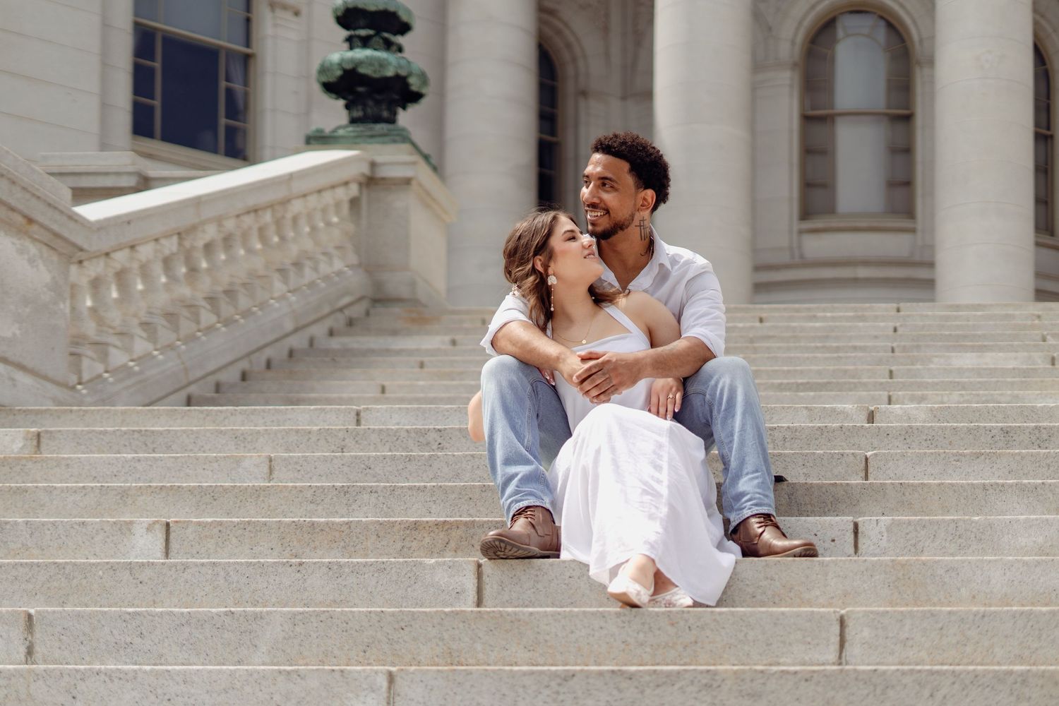 Couple sitting on stone steps of a grand building, sharing a romantic embrace on a sunny day.