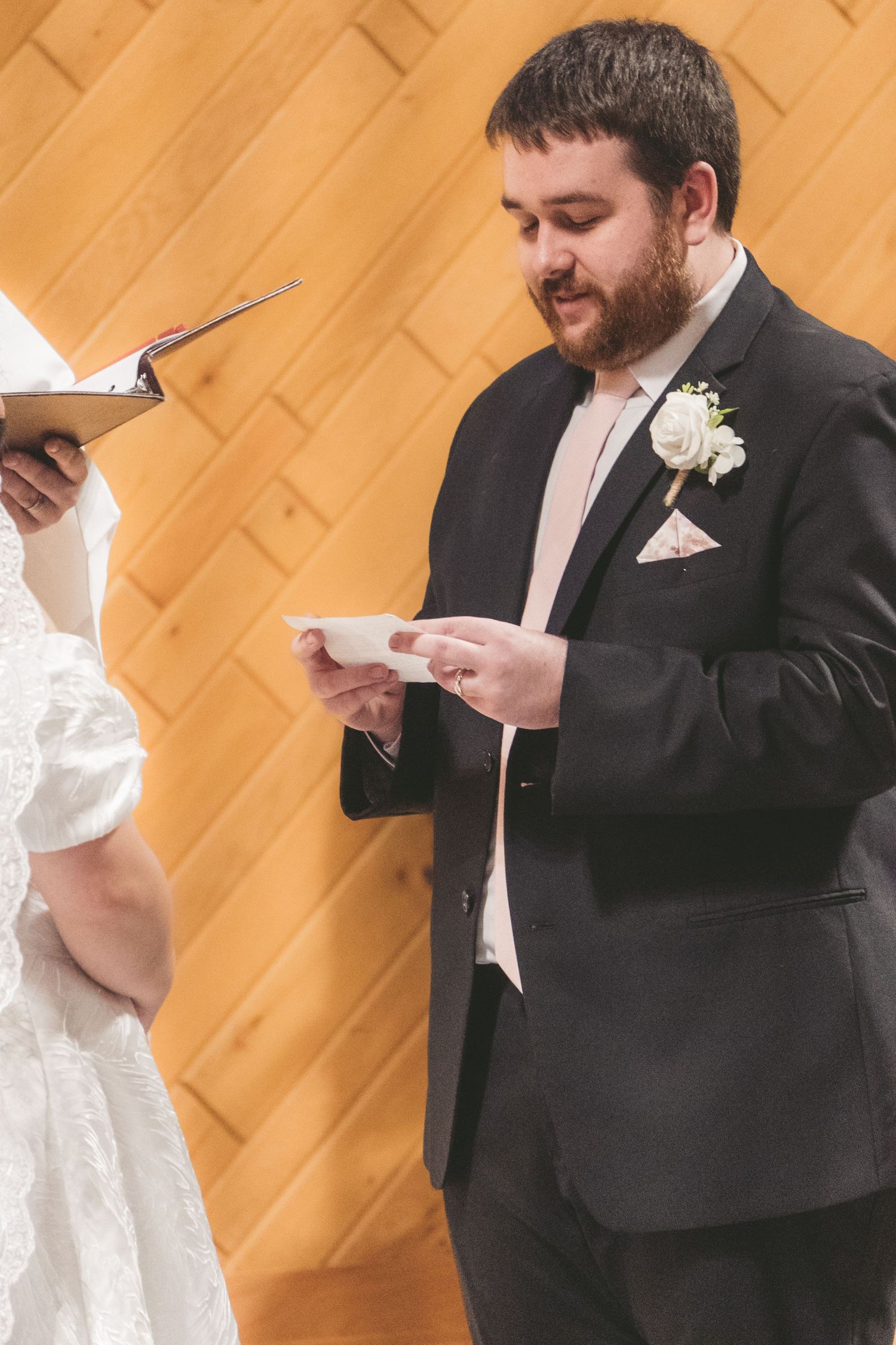 Groom in black suit reading wedding vows from a card during ceremony, white rose boutonniere, wooden wall background.