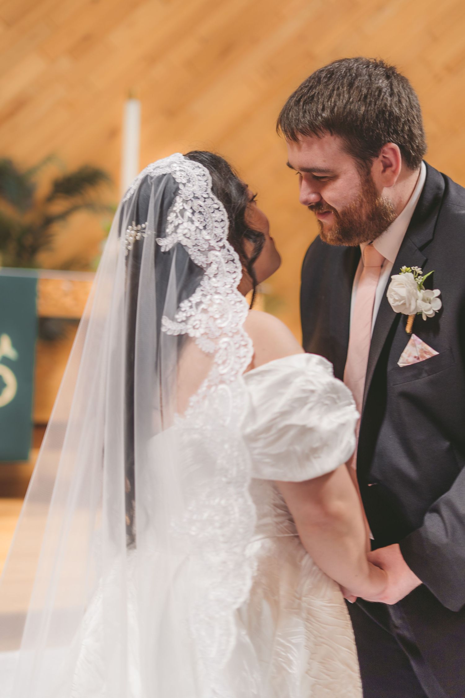 A bride in a lace veil and groom in a gray suit share an intimate moment at the altar during their wedding ceremony.