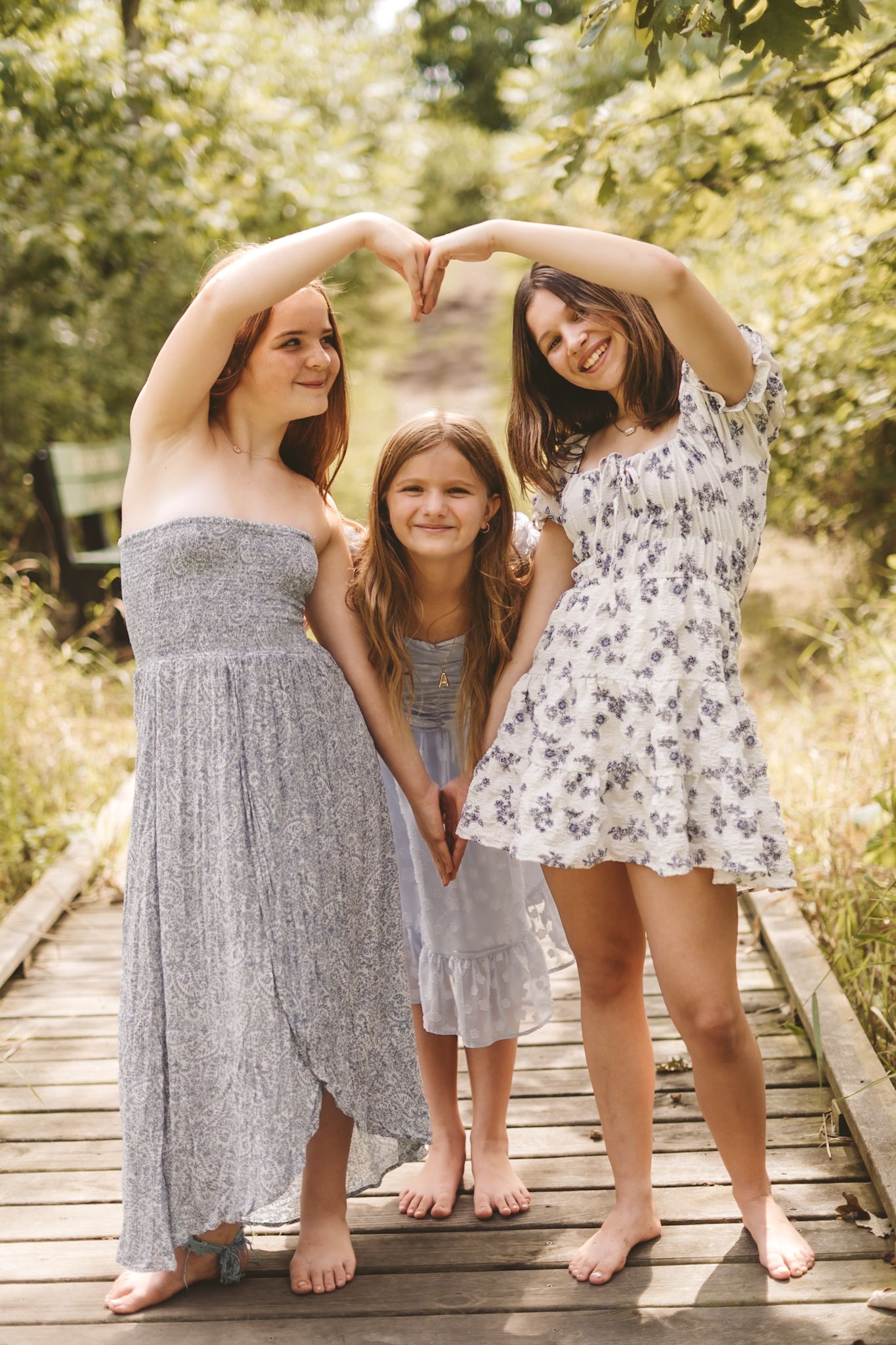 Three girls in summer dresses form a heart shape with their hands on a wooden boardwalk surrounded by green trees.