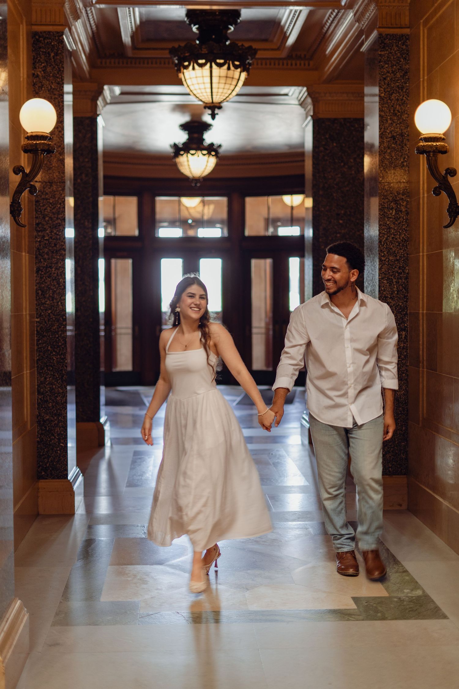 Happy couple holding hands walking through an elegant, warmly lit hallway with classic architecture.