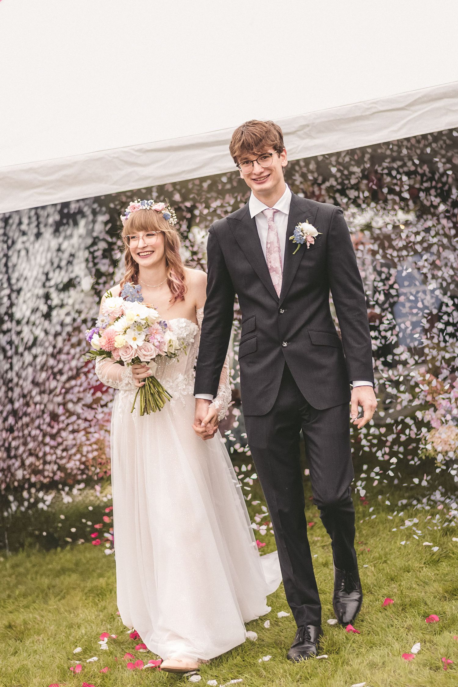 Newlyweds walking through confetti shower holding hands, bride carrying pink and white bouquet.