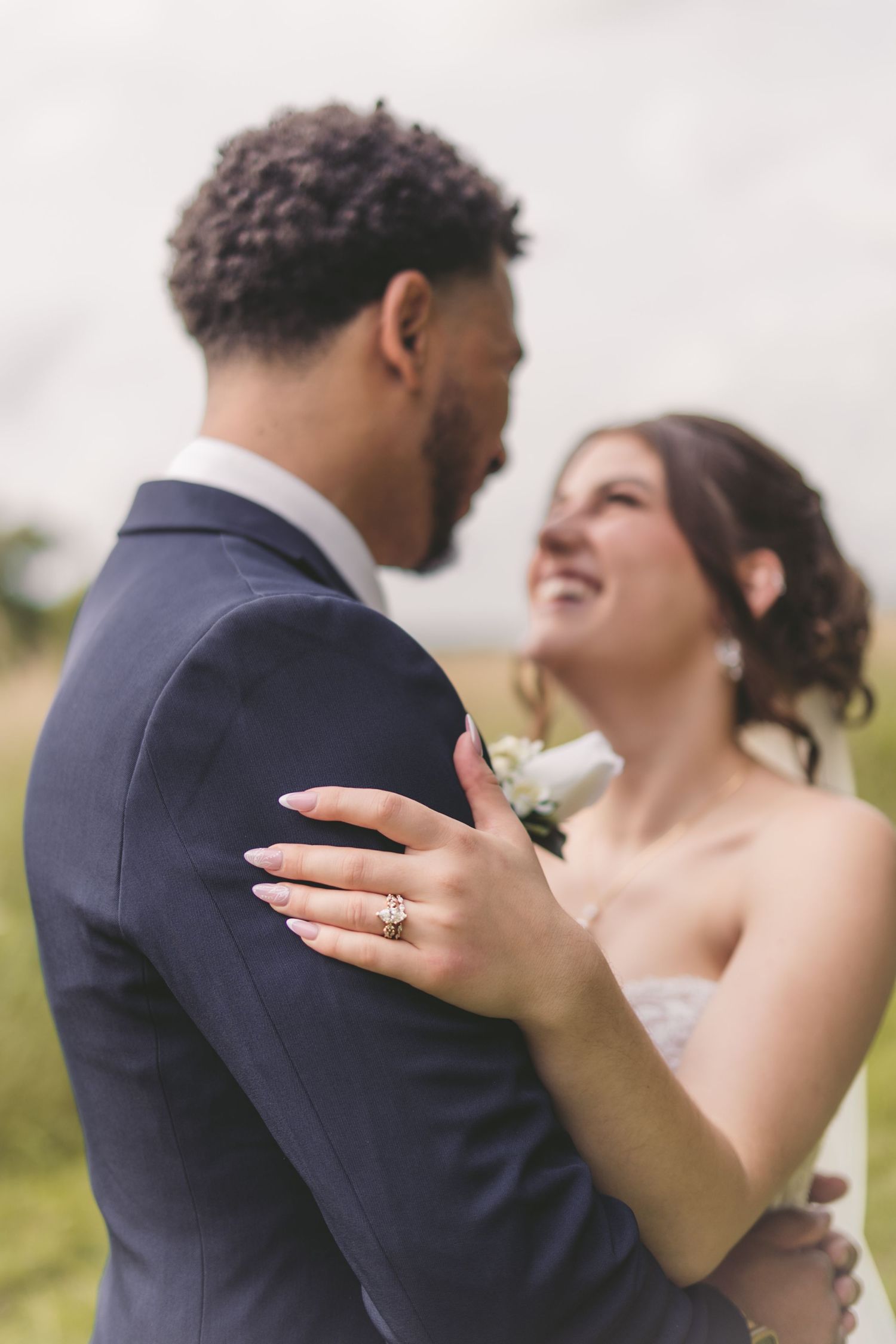 A smiling bride in a strapless gown embraces a groom in a navy suit, showcasing her diamond engagement ring.
