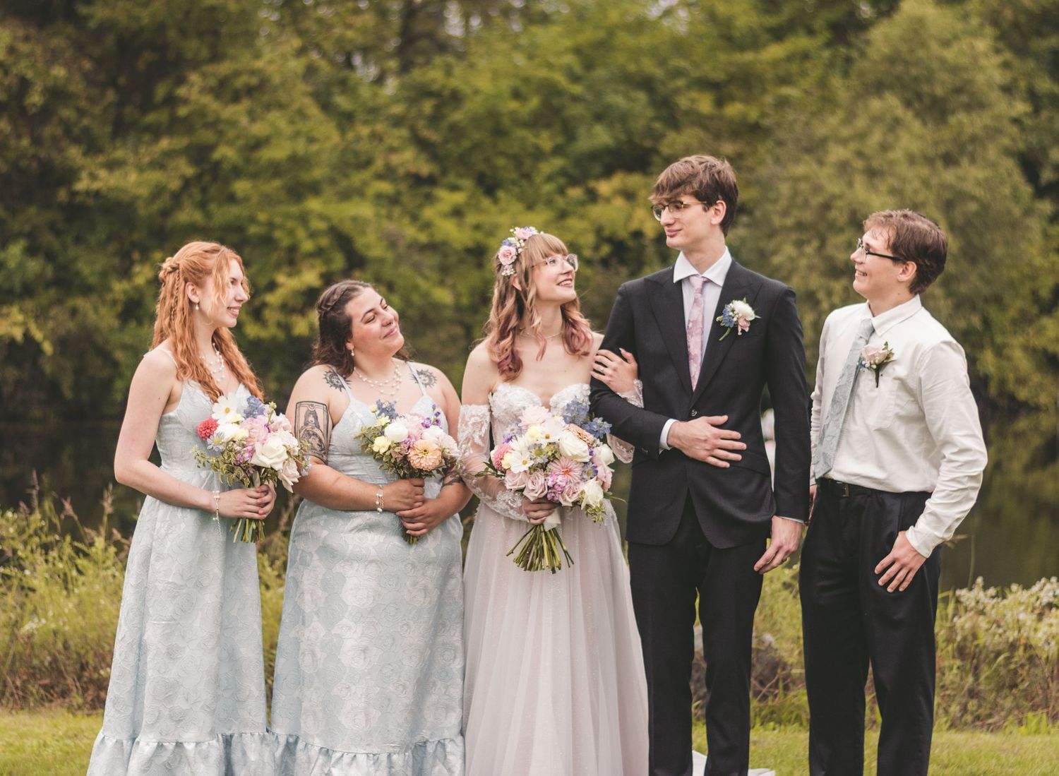 Wedding party with bride, groom, and bridesmaids in blue dresses posing outdoors near a pond.