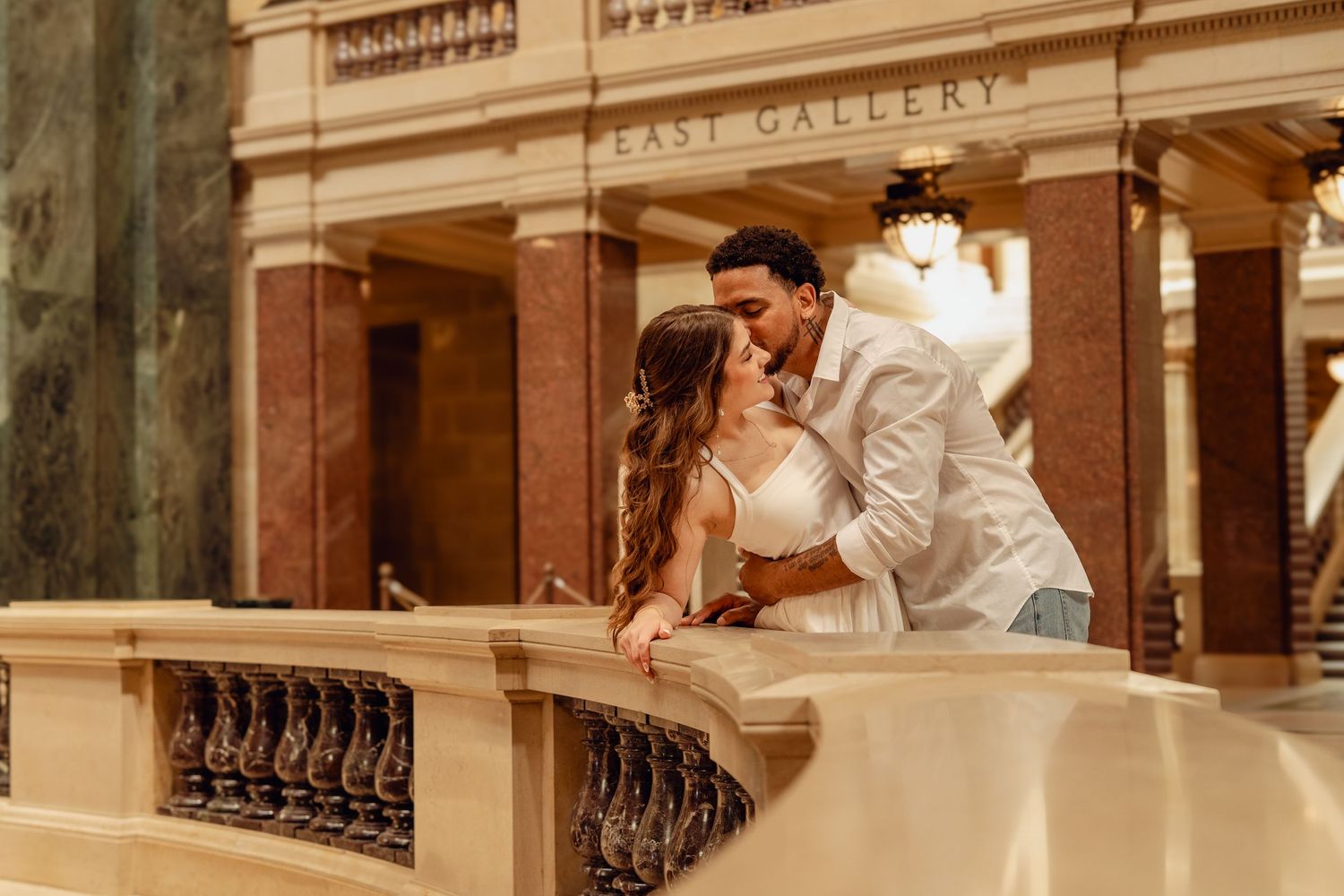 Interracial couple sharing a kiss on ornate balcony inside East Gallery of elegant historic building.