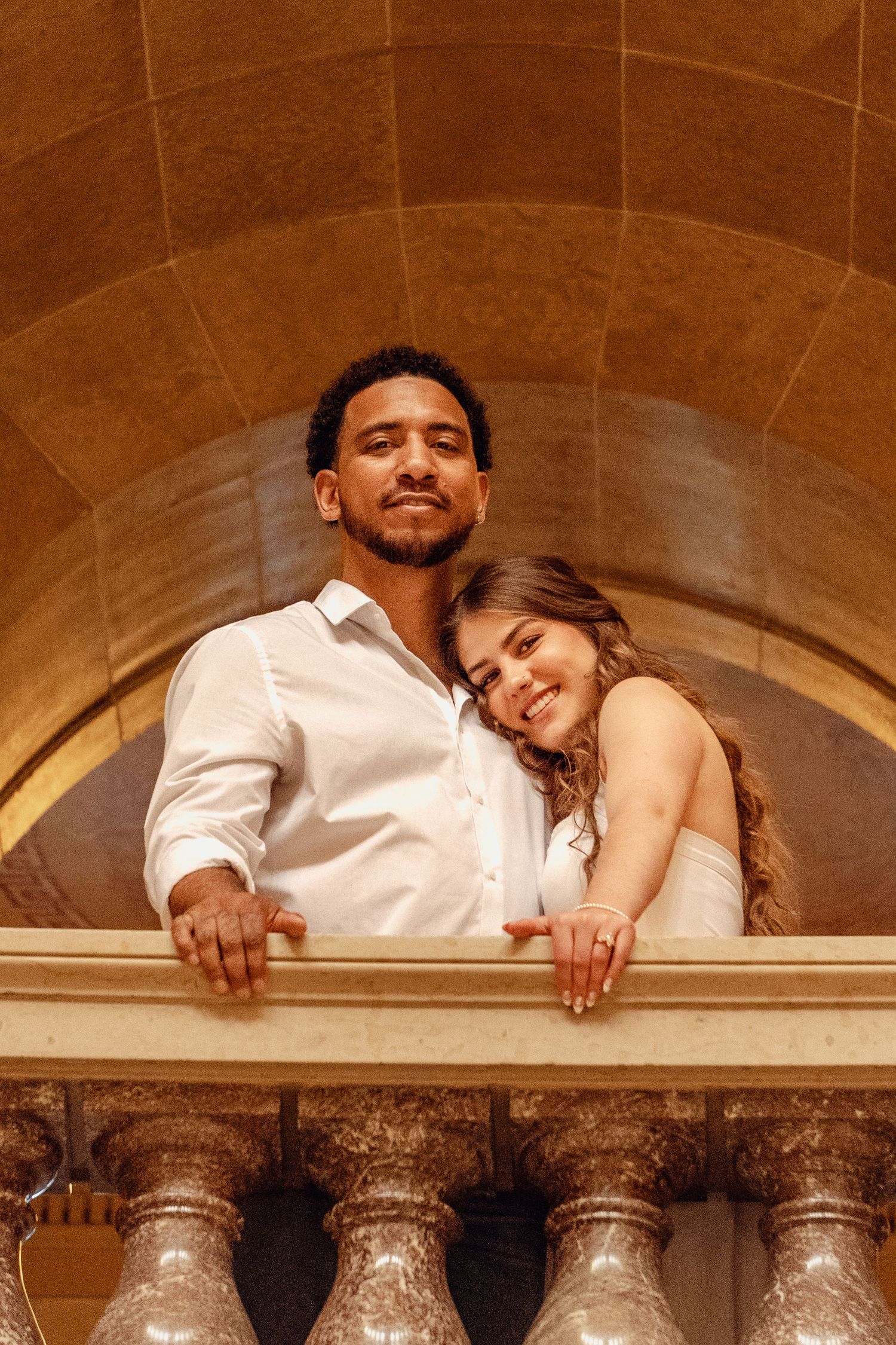 Romantic couple posing on an ornate balcony with stone arches, woman leaning on man in white shirt.