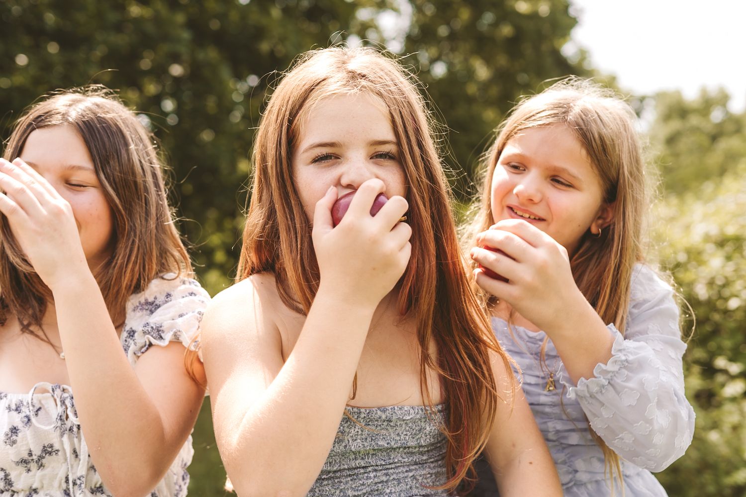 Three girls eating fresh apples outdoors in a sunny garden, smiling and enjoying a healthy snack together.