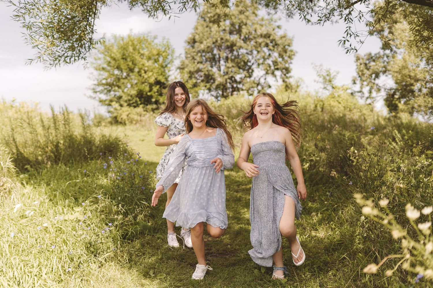Three girls in matching gray dresses run joyfully through a lush green meadow path on a sunny summer day.