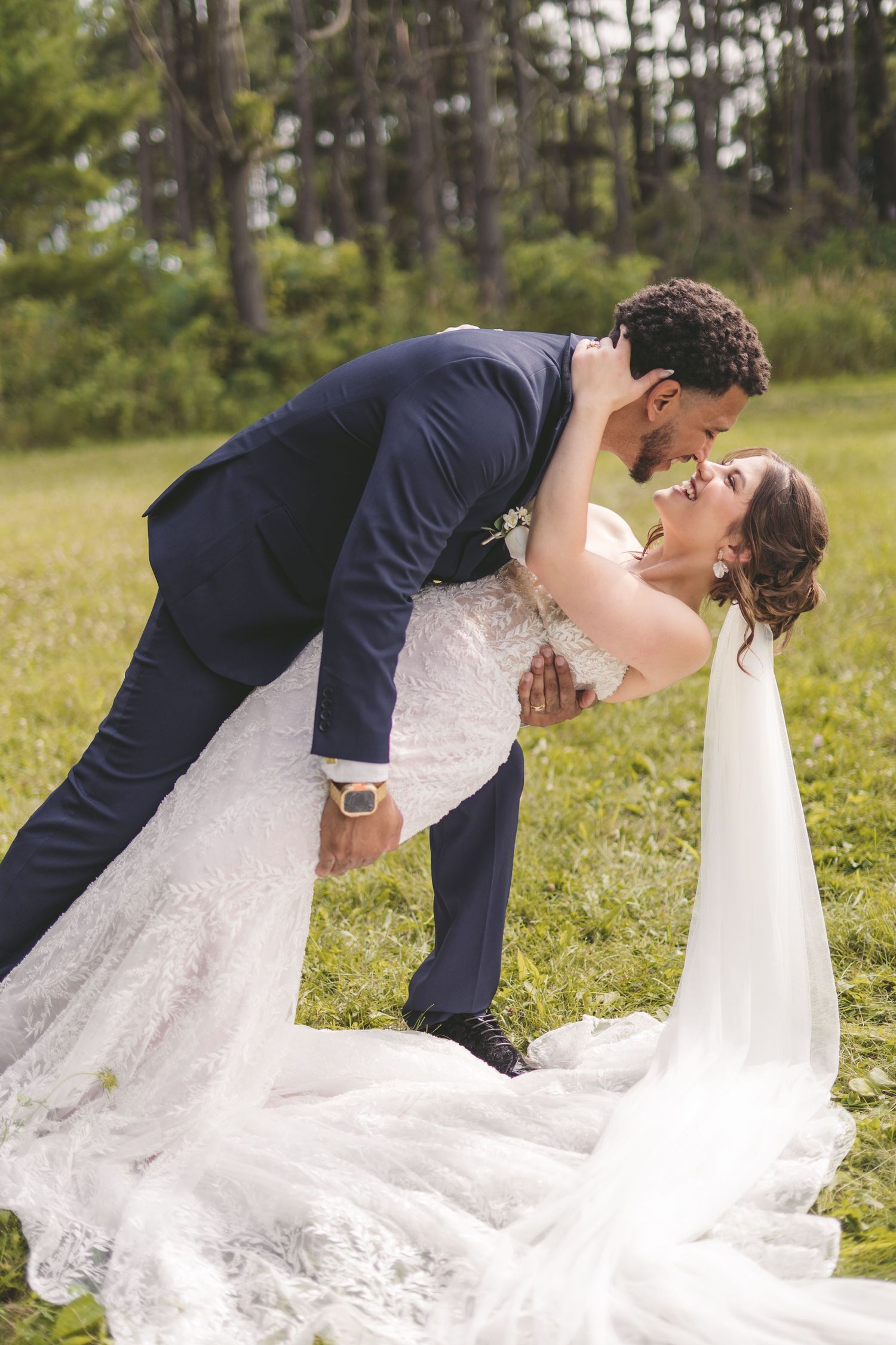 Groom in navy suit dips bride in white lace gown for a romantic kiss on a green lawn with trees.