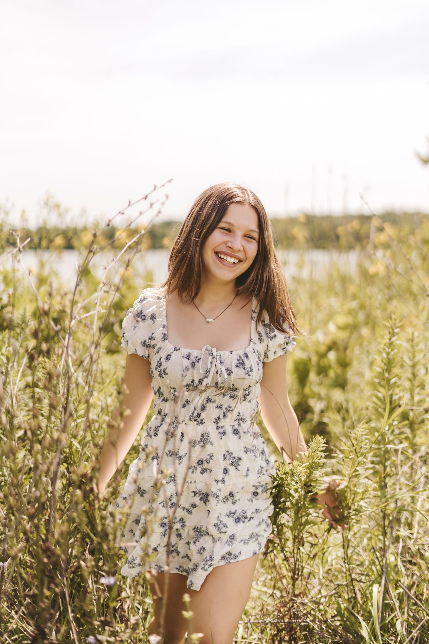 Smiling teenage girl in floral sundress walking through wildflower field on sunny day.