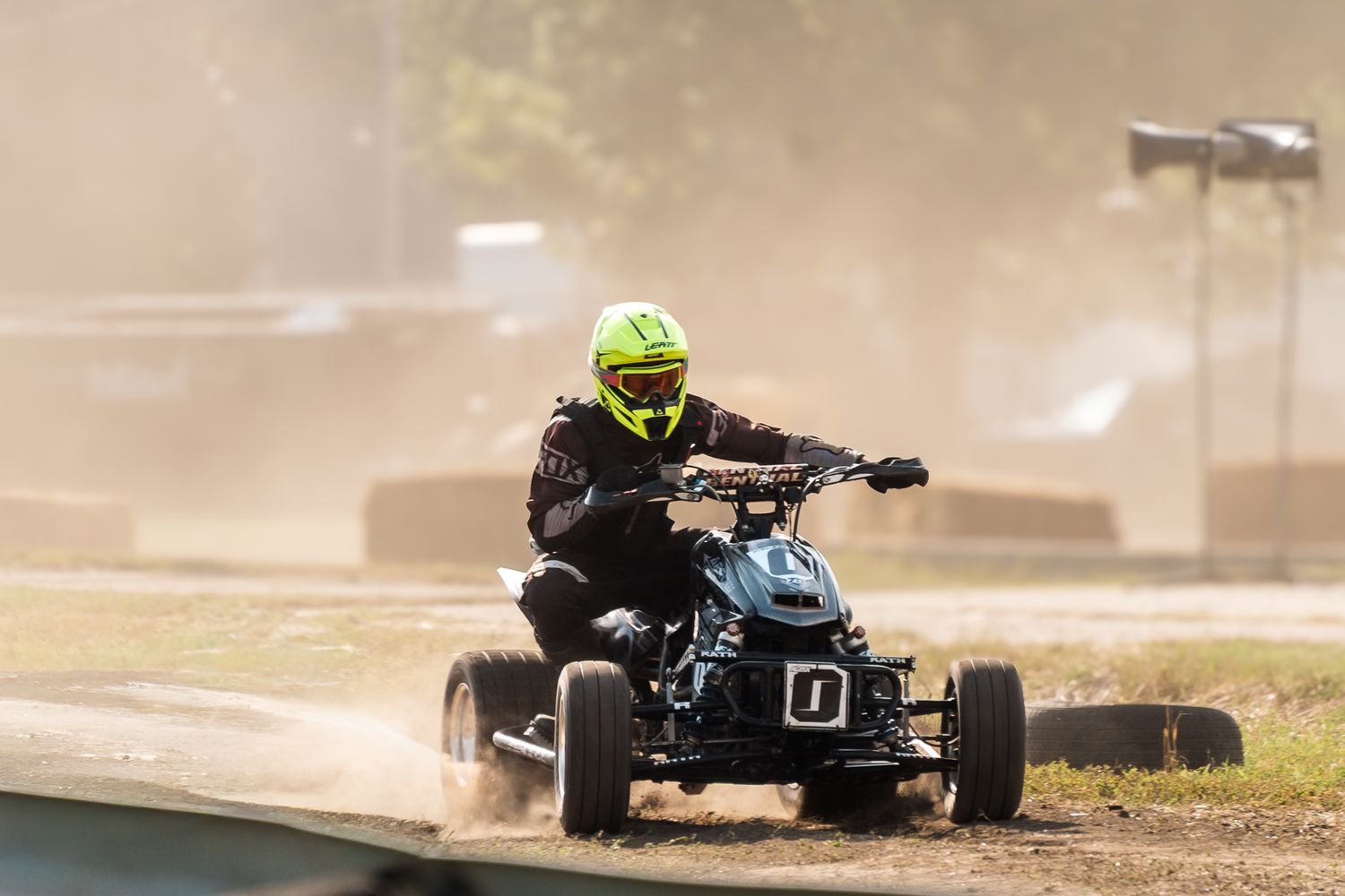 A rider in a yellow helmet races a quad bike on a dusty dirt track, kicking up clouds of dust.