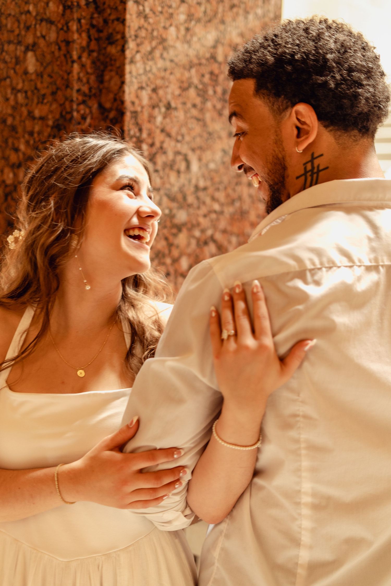A joyful interracial couple embracing and smiling at each other outdoors, dressed in elegant cream attire.