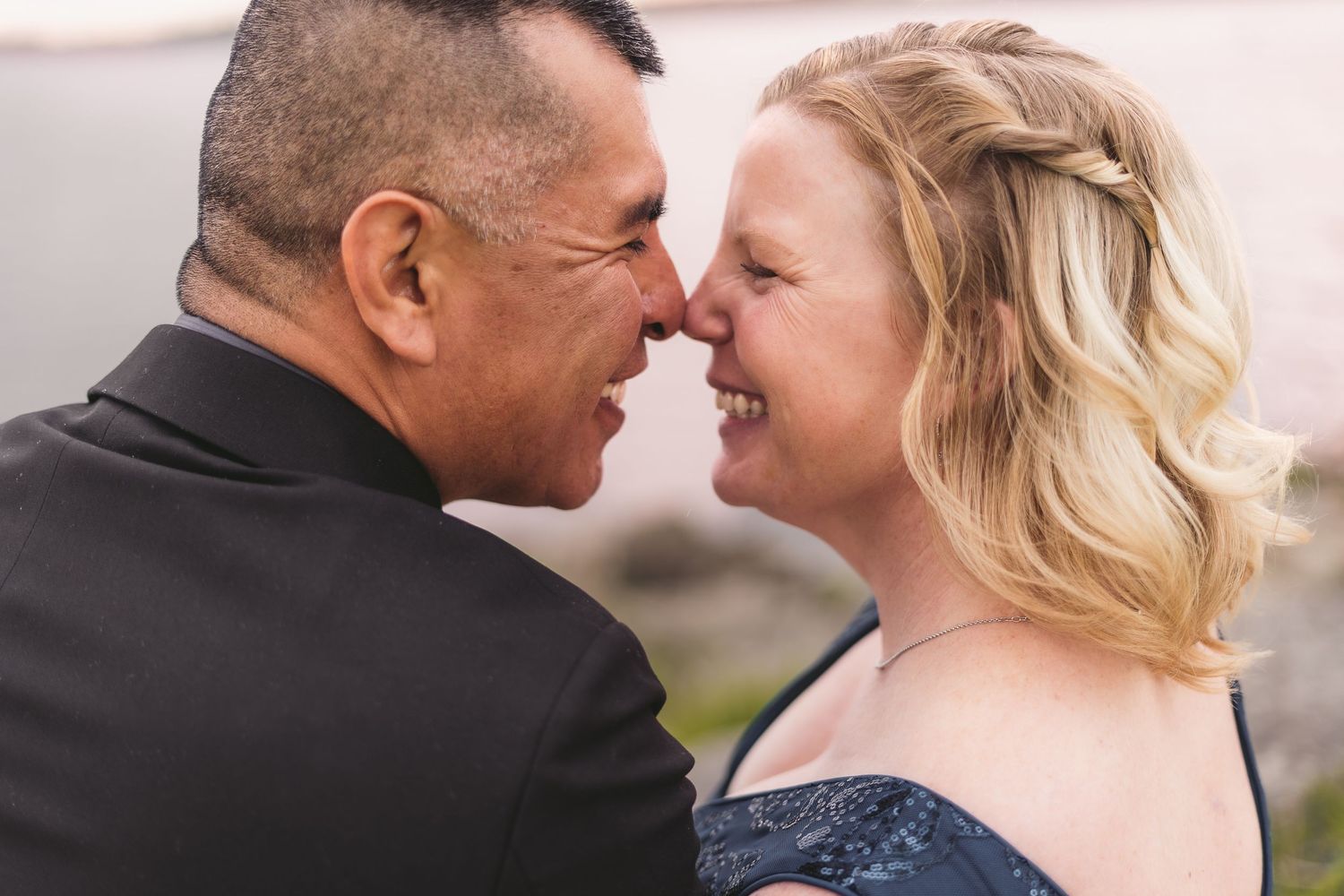 A joyful couple sharing an intimate moment, touching noses and smiling, dressed in formal attire at an outdoor wedding.