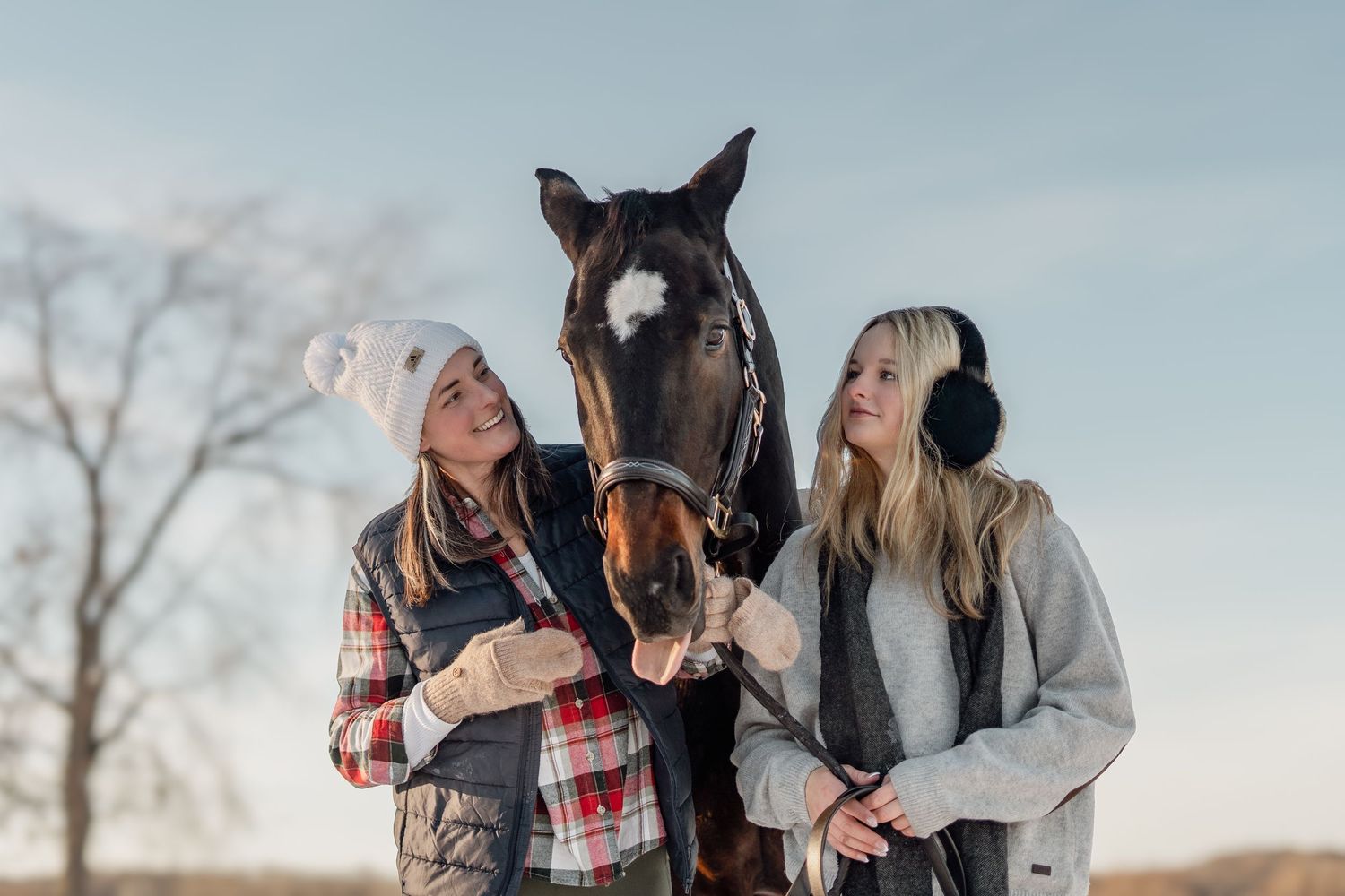 Two young women in winter attire stand with a dark horse outdoors on a cold day.