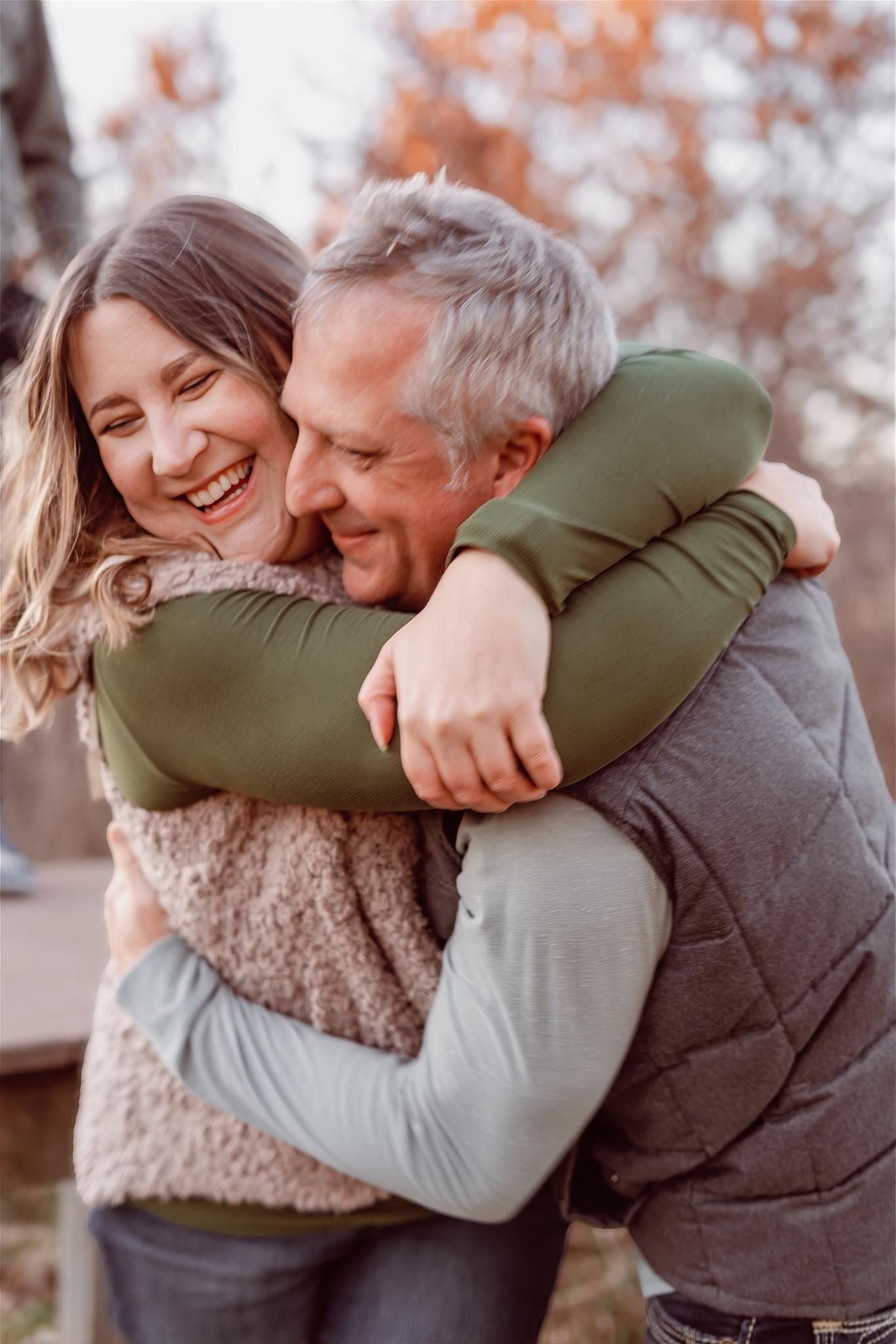 A laughing woman and smiling older man share a warm embrace outdoors during autumn.
