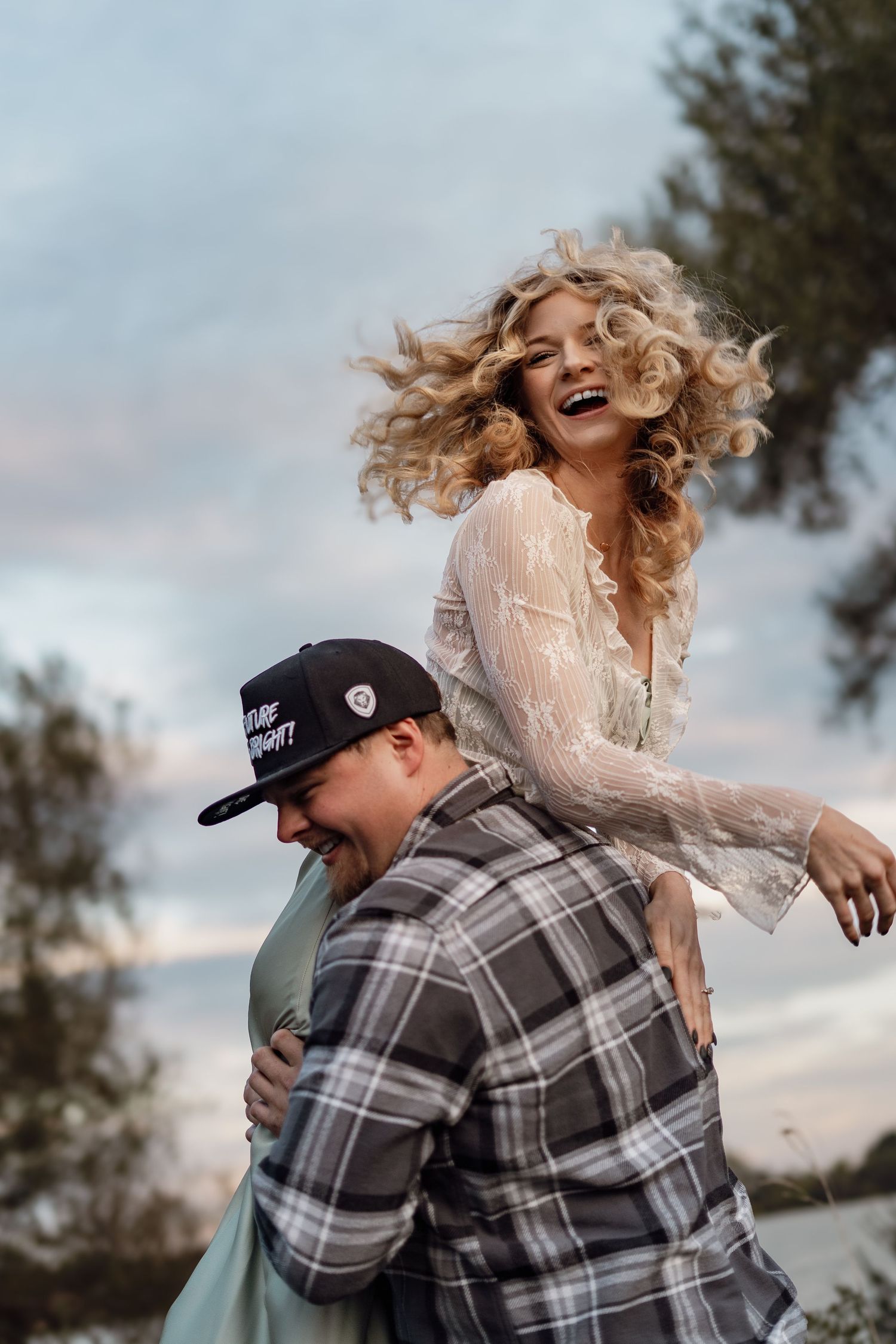 Playful couple embracing and laughing in casual attire against a cloudy sky.