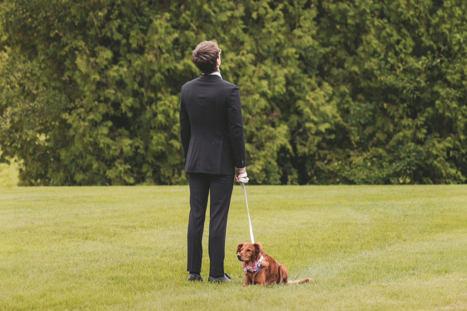 Man in black suit standing on green lawn holding leash with small brown dog sitting beside him.