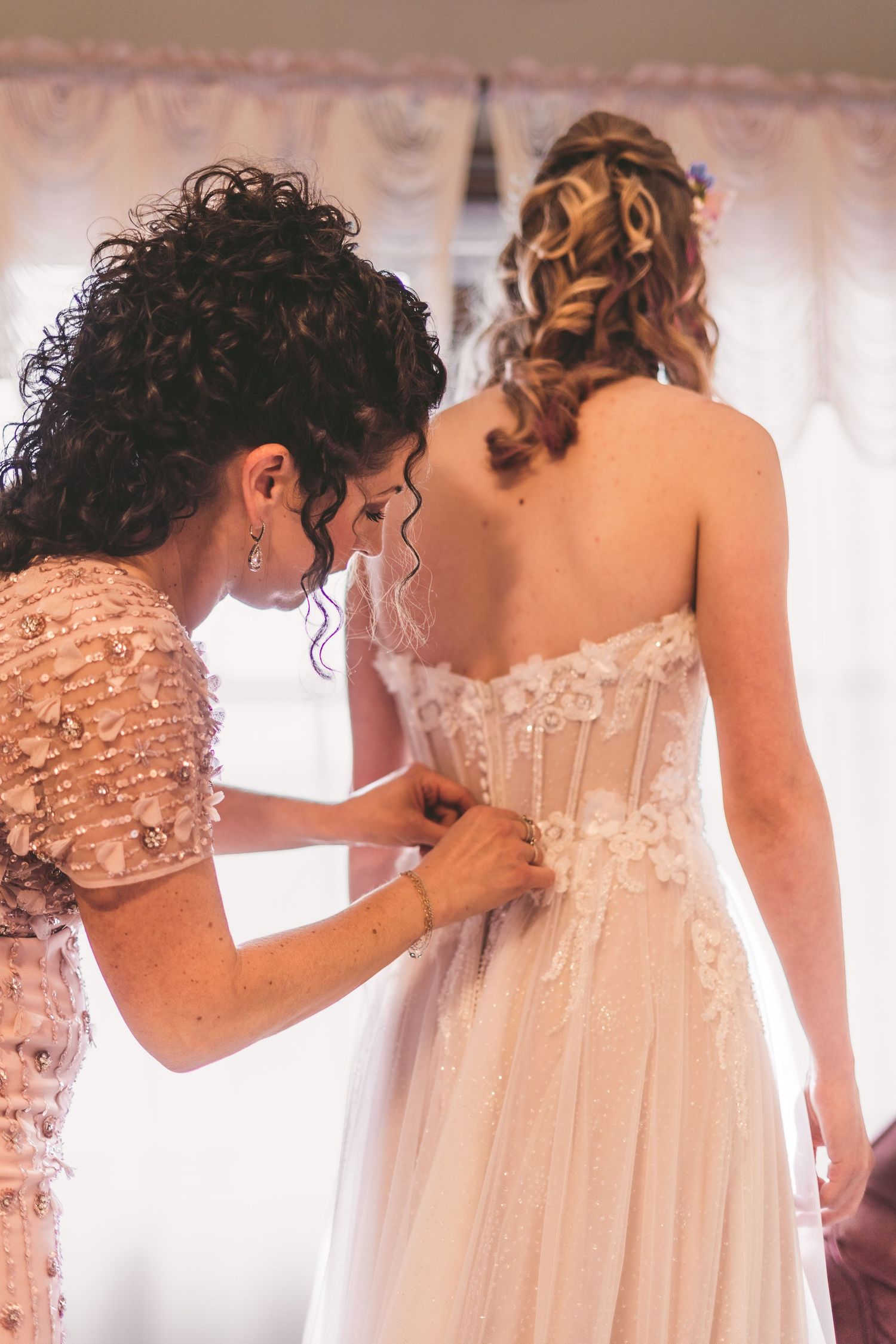 A bridesmaid helps button the back of a brides floral lace wedding gown during bridal preparations.