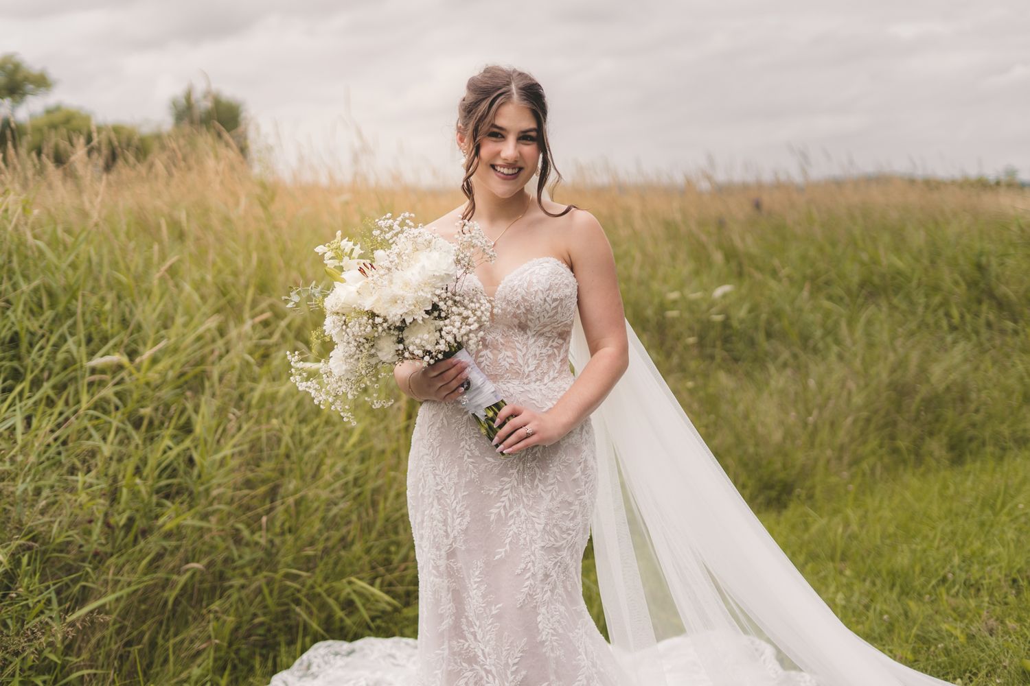 Bride in lace mermaid gown holding white bouquet standing in green field on cloudy day.