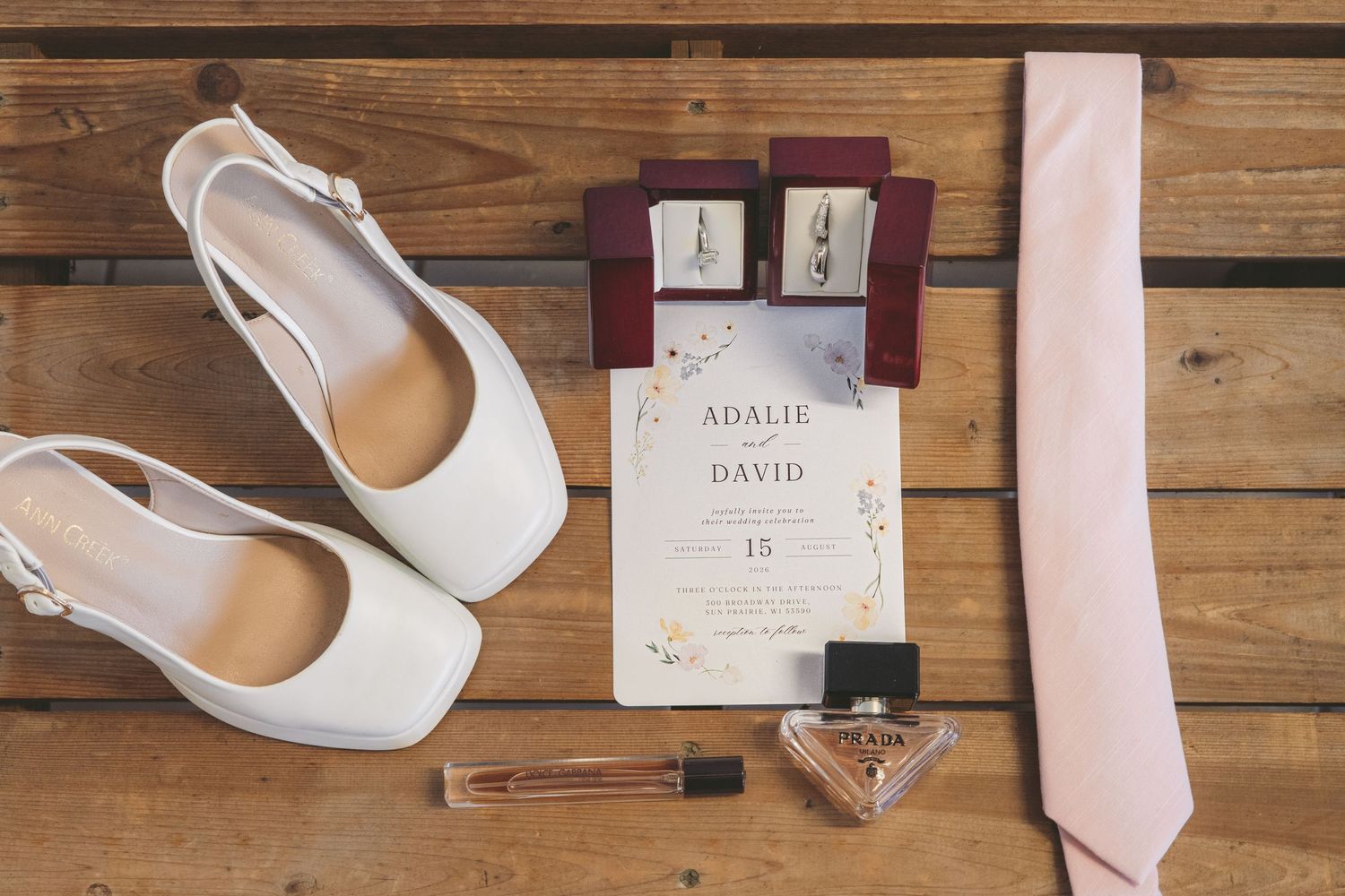 Wedding flat lay featuring white bridal heels, pink tie, floral invitation for Adalie and David, earrings, and perfume on wood.