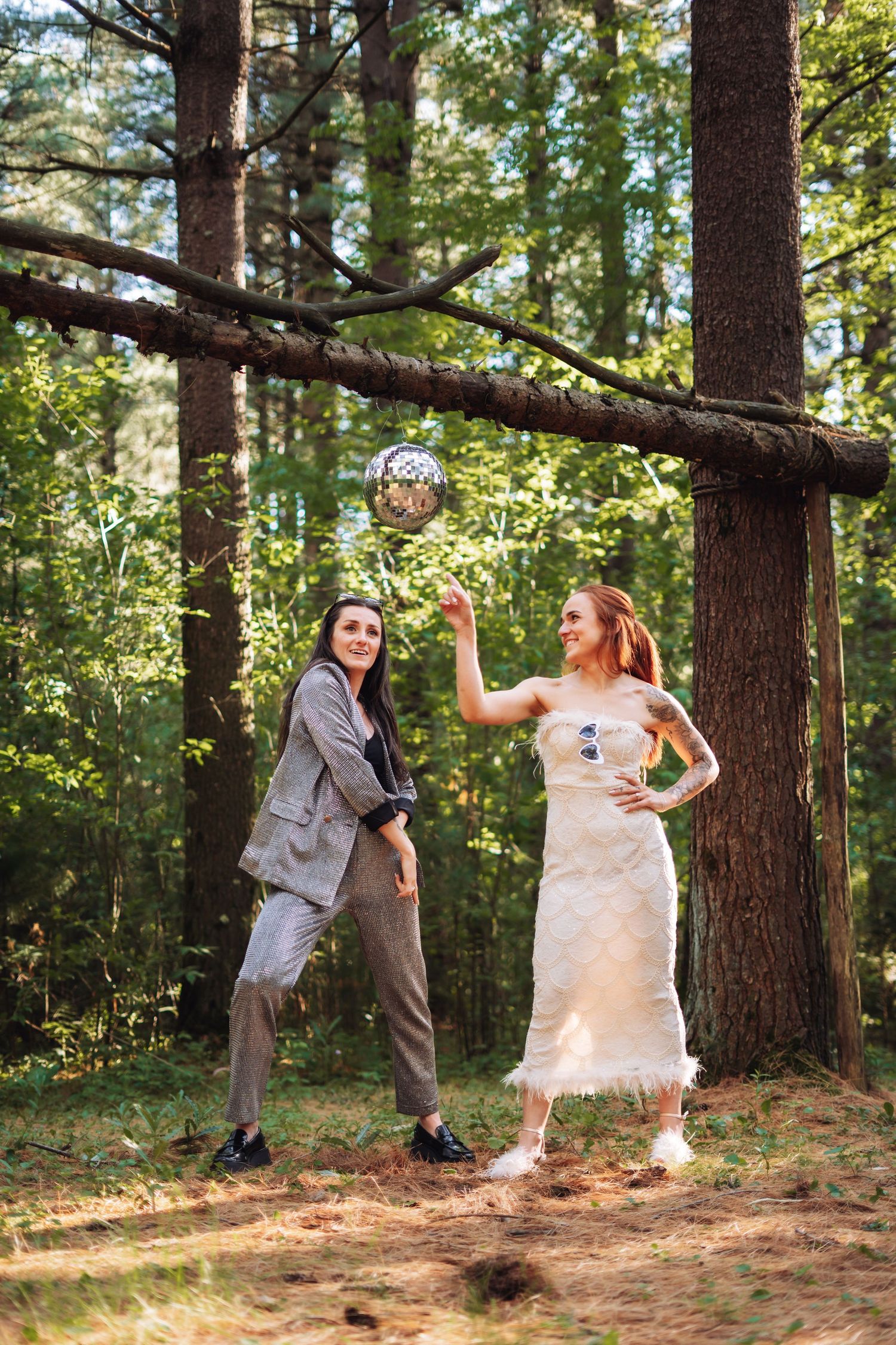 Two women pose playfully in a forest, one in a suit and one in a white dress, holding a disco ball ornament.