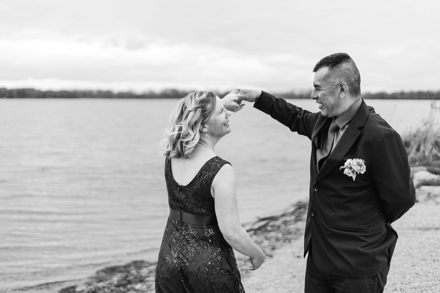 Black and white photo of a couple dancing by a lakeside, groom in suit with boutonniere, bride in elegant dress.